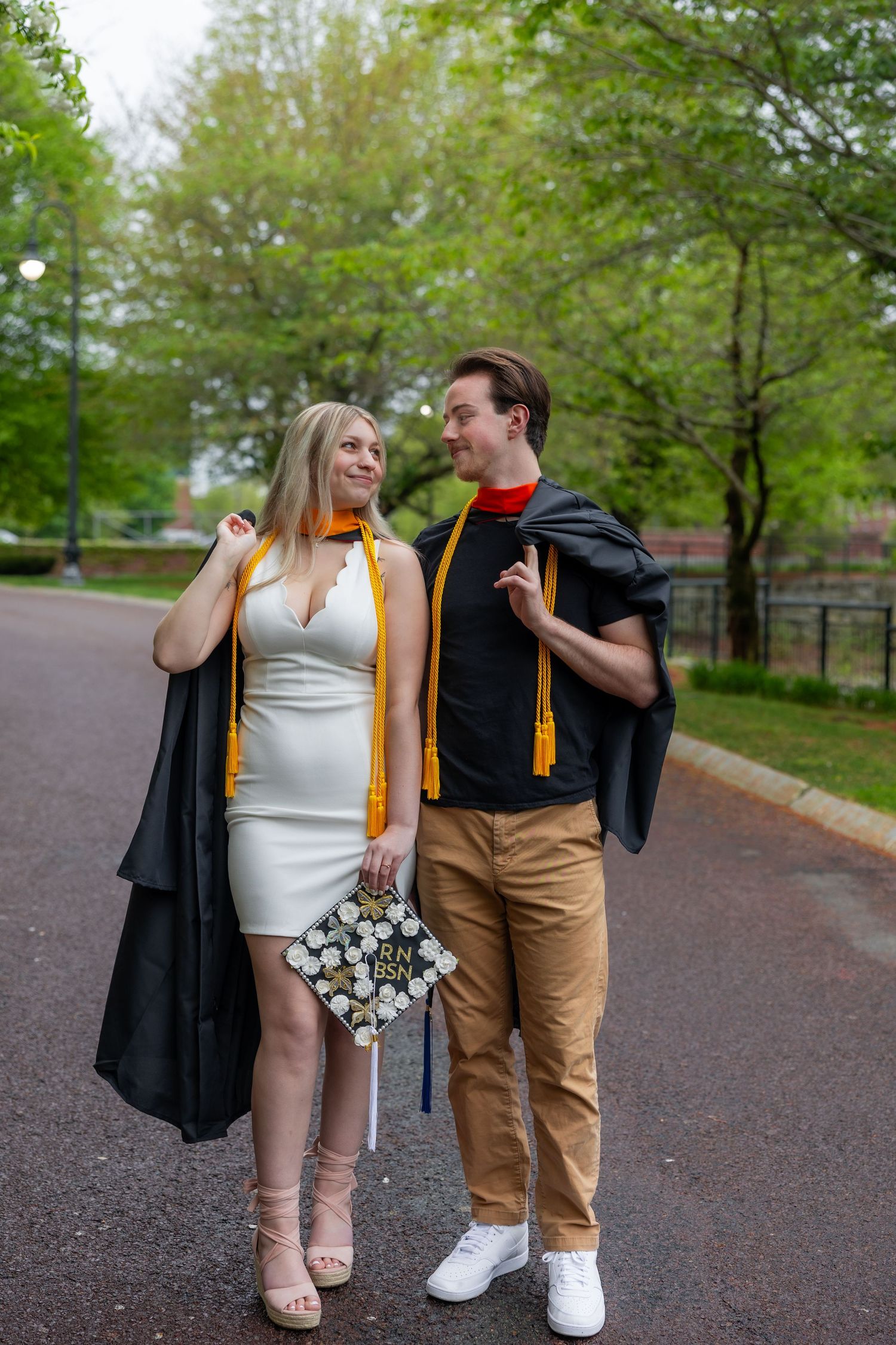 A couple in graduation attire walks together on a tree-lined path holding a bouquet of flowers.