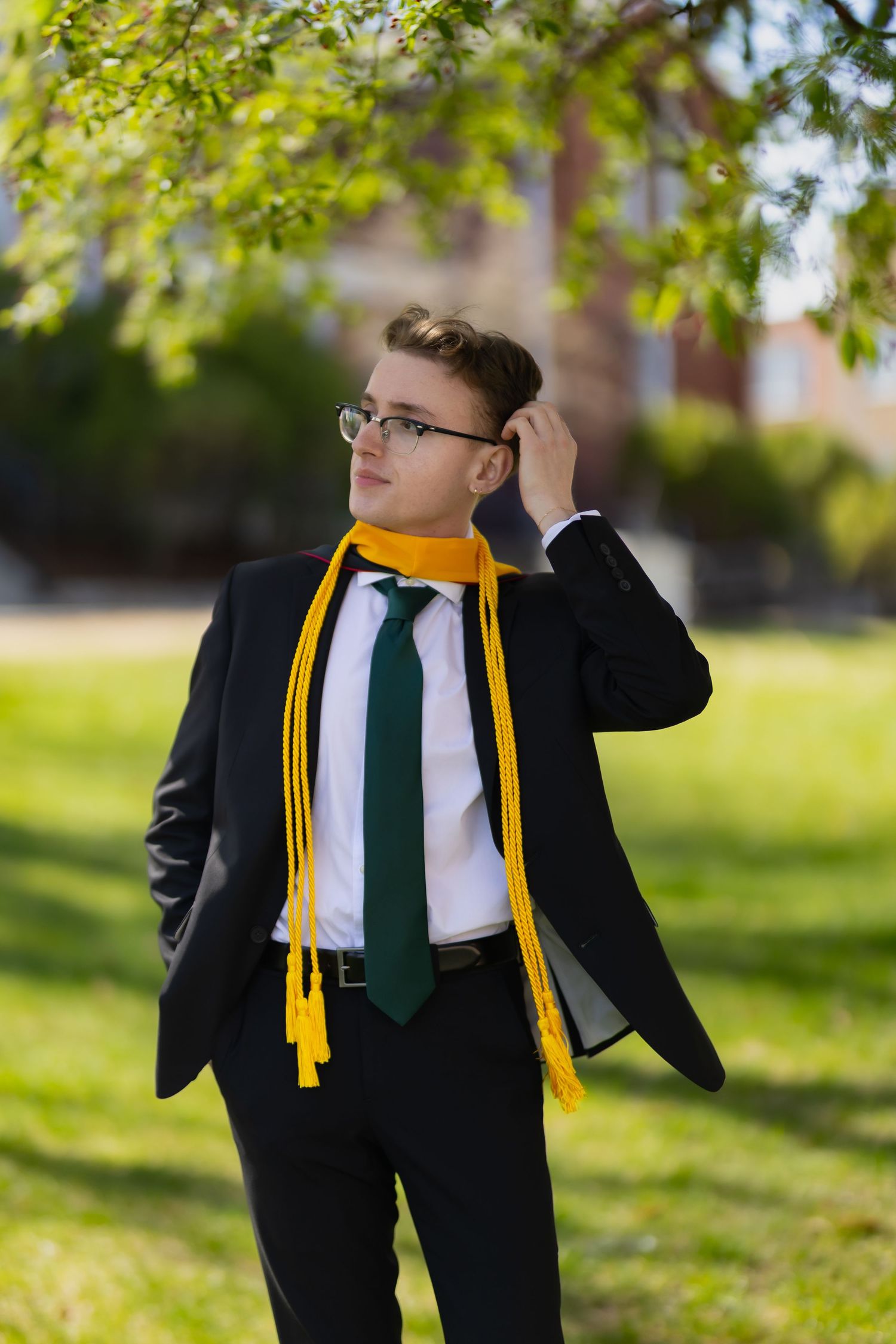Young man in black suit, green tie, and yellow graduation cords standing outdoors on a sunny day.