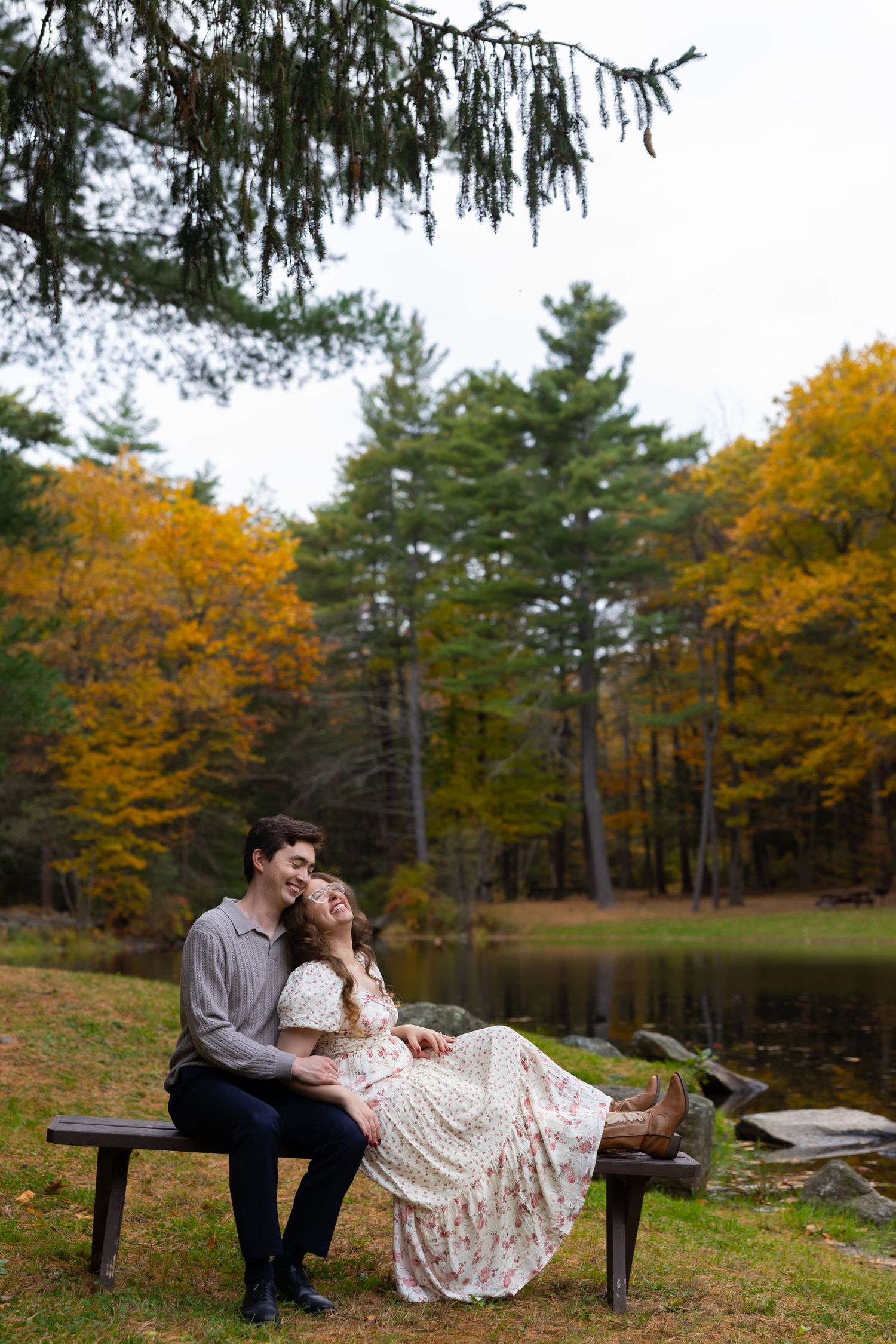 Couple sitting on a bench by a pond in an autumn forest, woman in floral dress laughing with man in gray shirt.