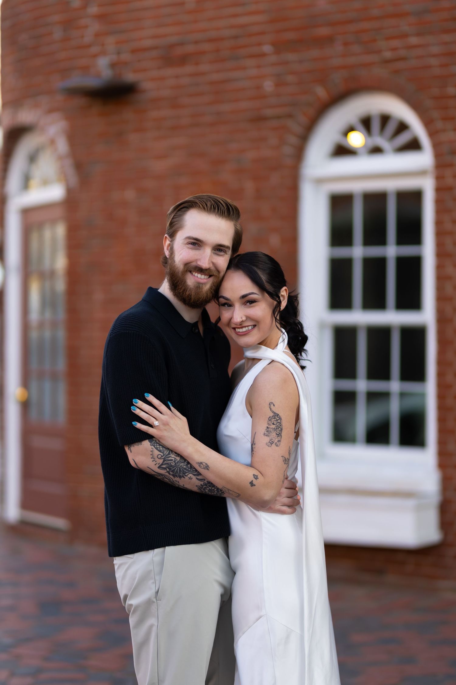 A couple embraces and smiles in front of a brick building, the woman in white and the man in black, both with tattoos.