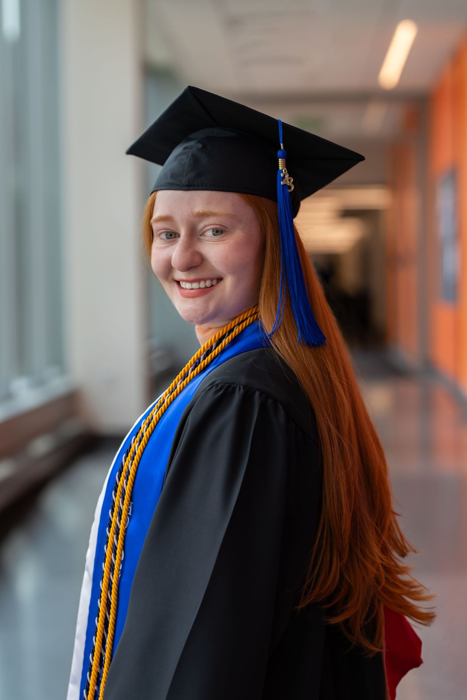 Graduate wearing traditional black cap and gown with blue and gold honor cords smiles in hallway.