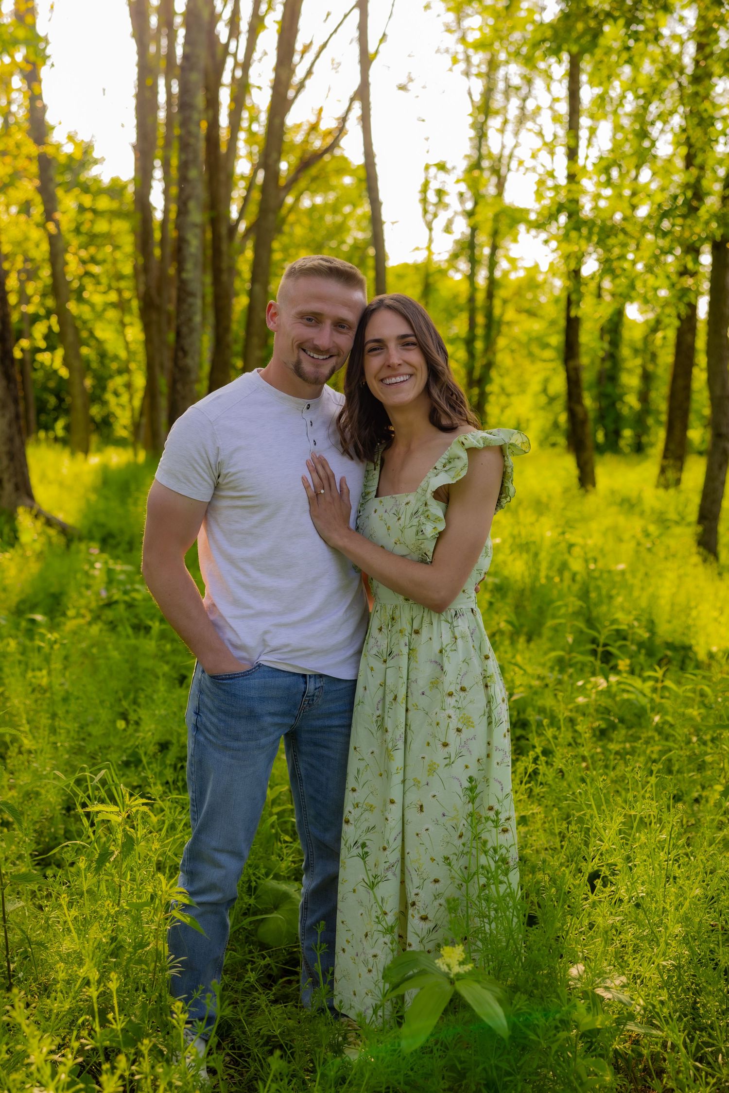 A couple poses together in a sunlit forest with green grass and trees during golden hour.