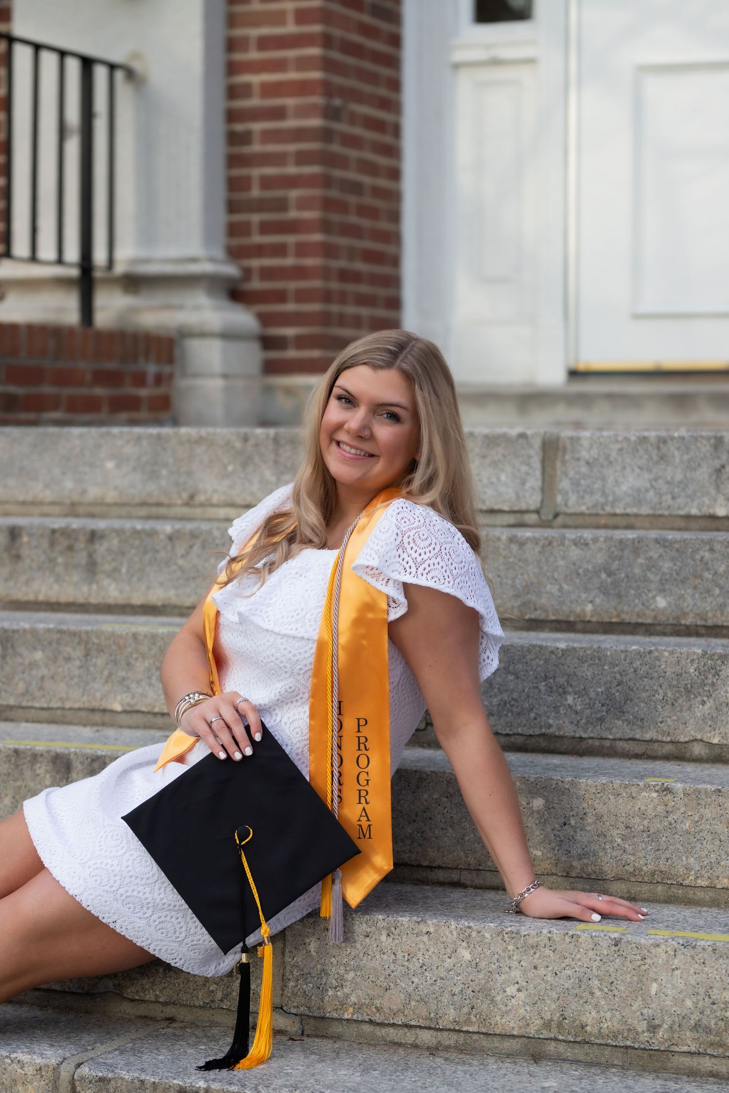 A person in a white eyelet dress and gold graduation cap poses on stone steps in a collage of four similar poses.
