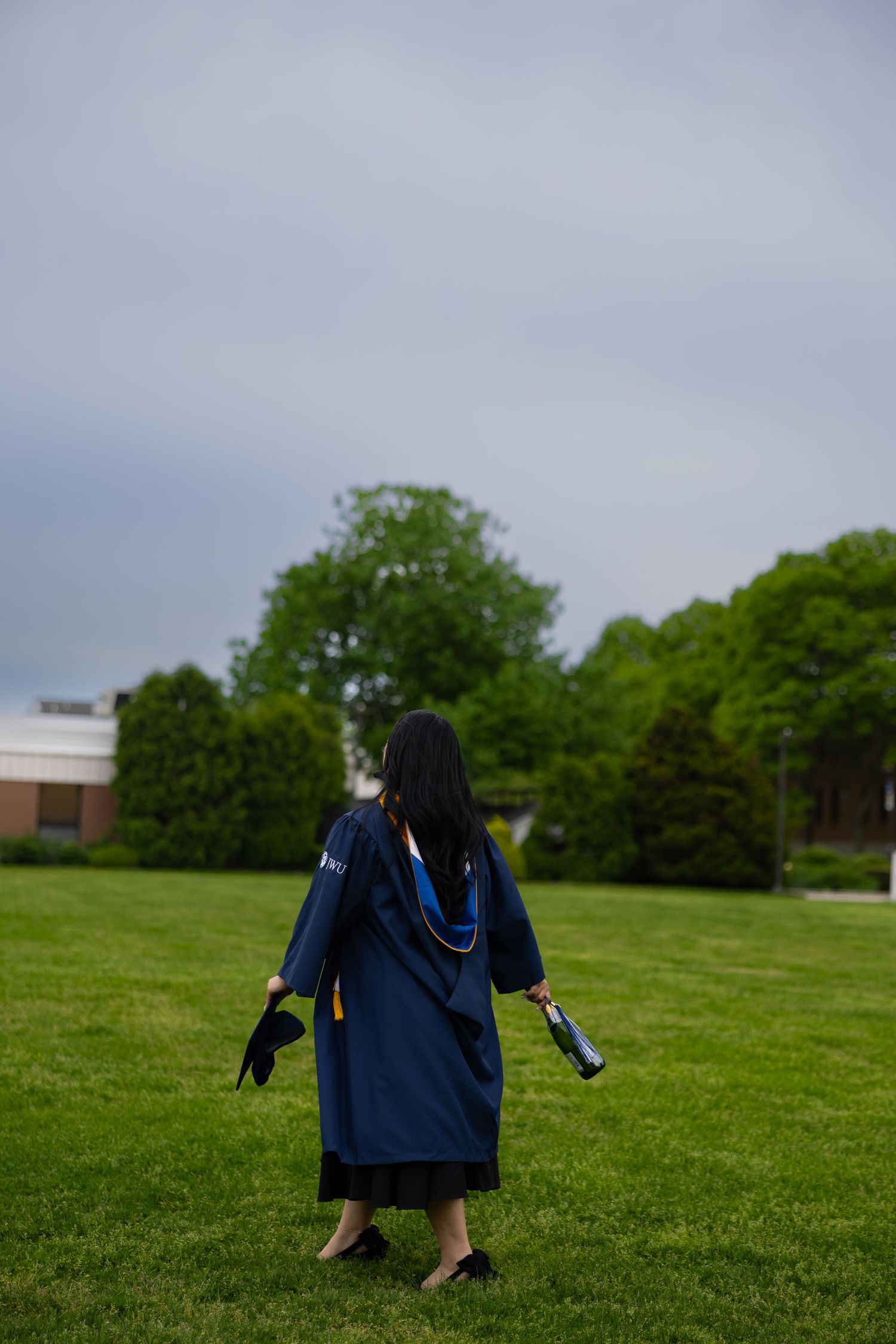 Graduate in navy cap and gown walks across green campus lawn, holding diploma and cap on cloudy day.