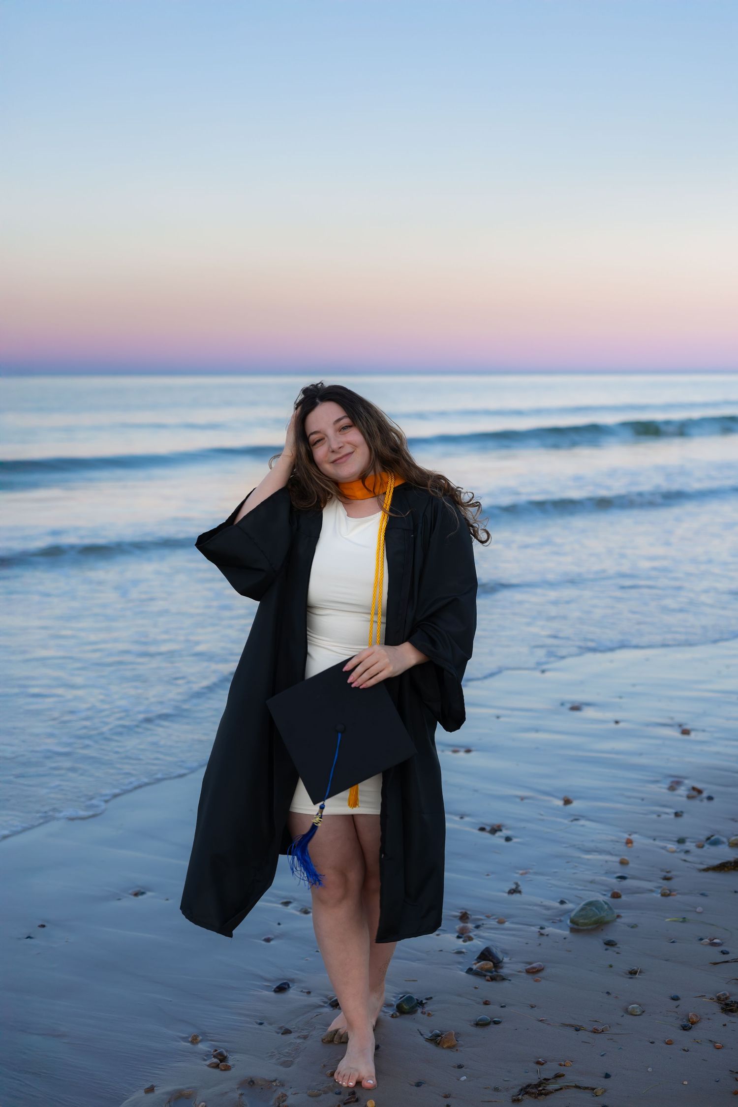 Person in black coat and white top poses playfully on beach at sunset with ocean waves and scattered shells in background.