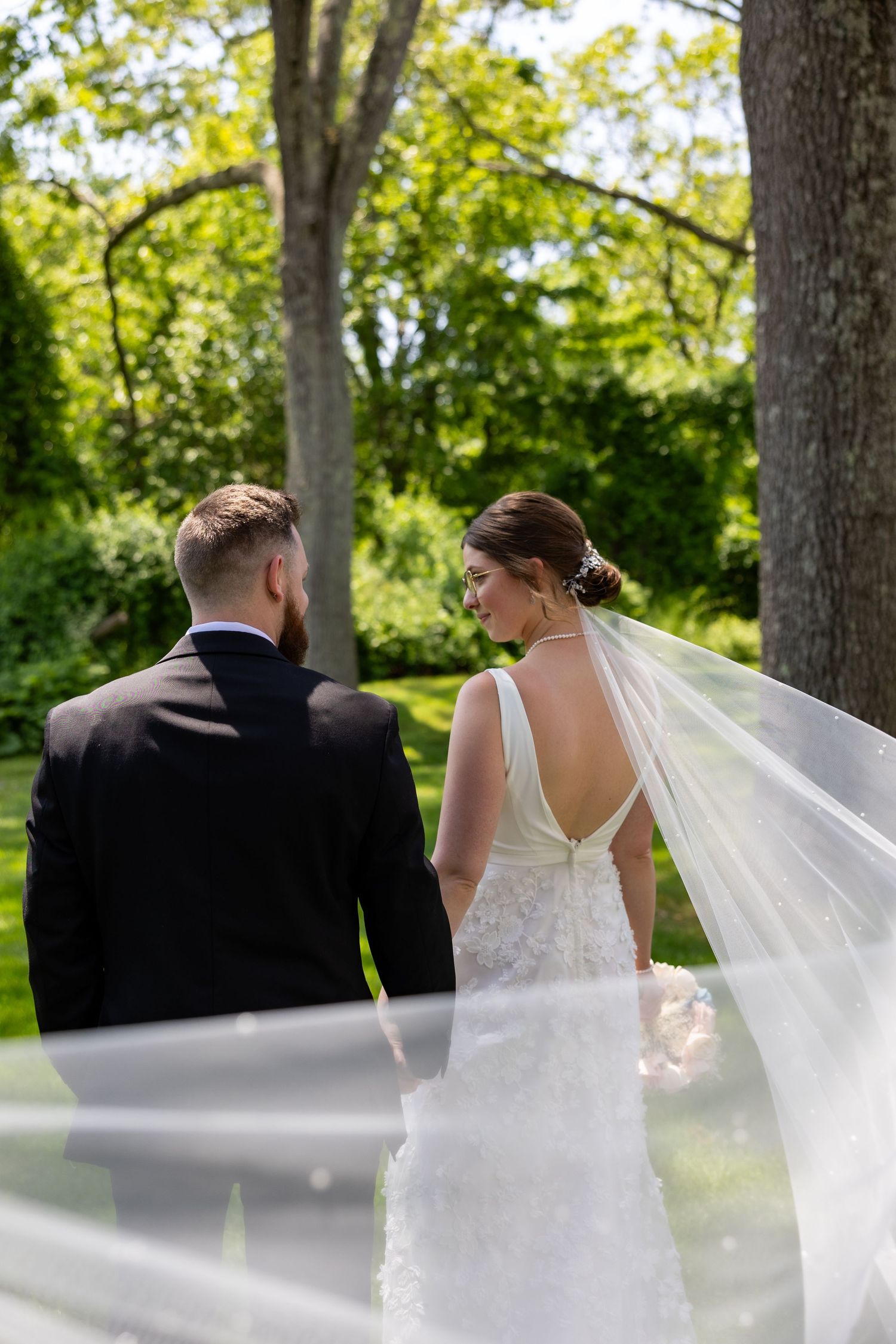 Elegant wedding couple walks through a sunlit garden as the bride's long veil flows gracefully in the breeze.