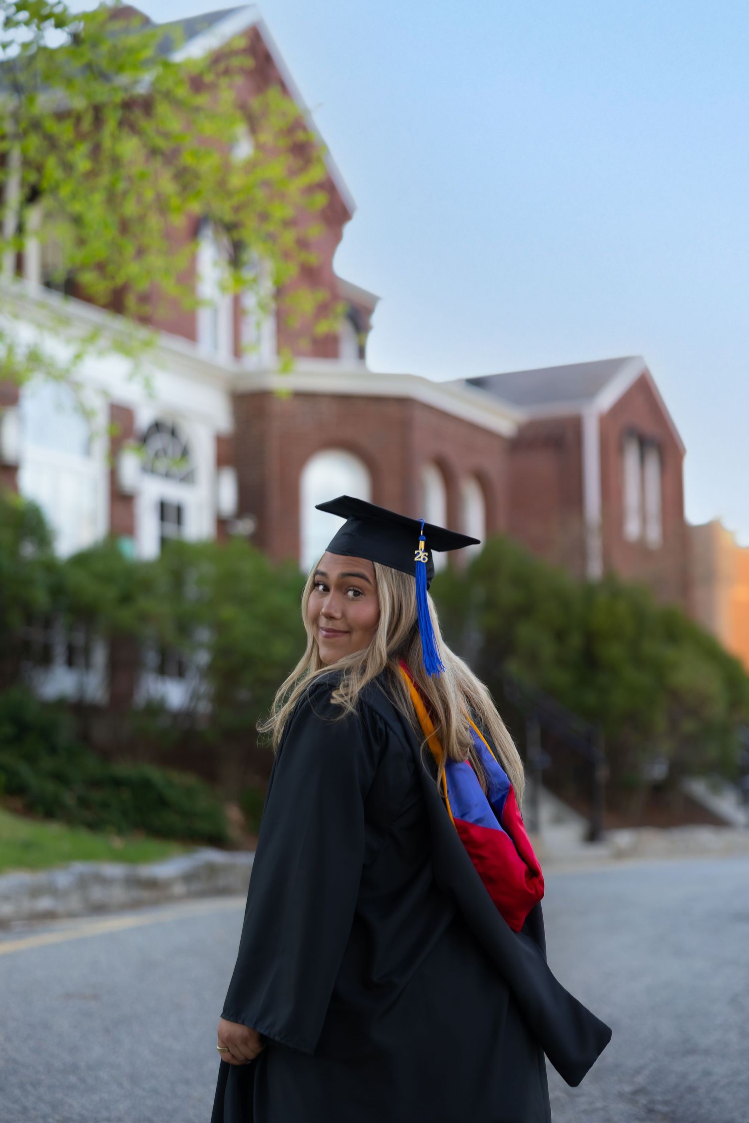 A smiling female graduate in black cap and gown poses on a brick campus path with a red brick building in the background.