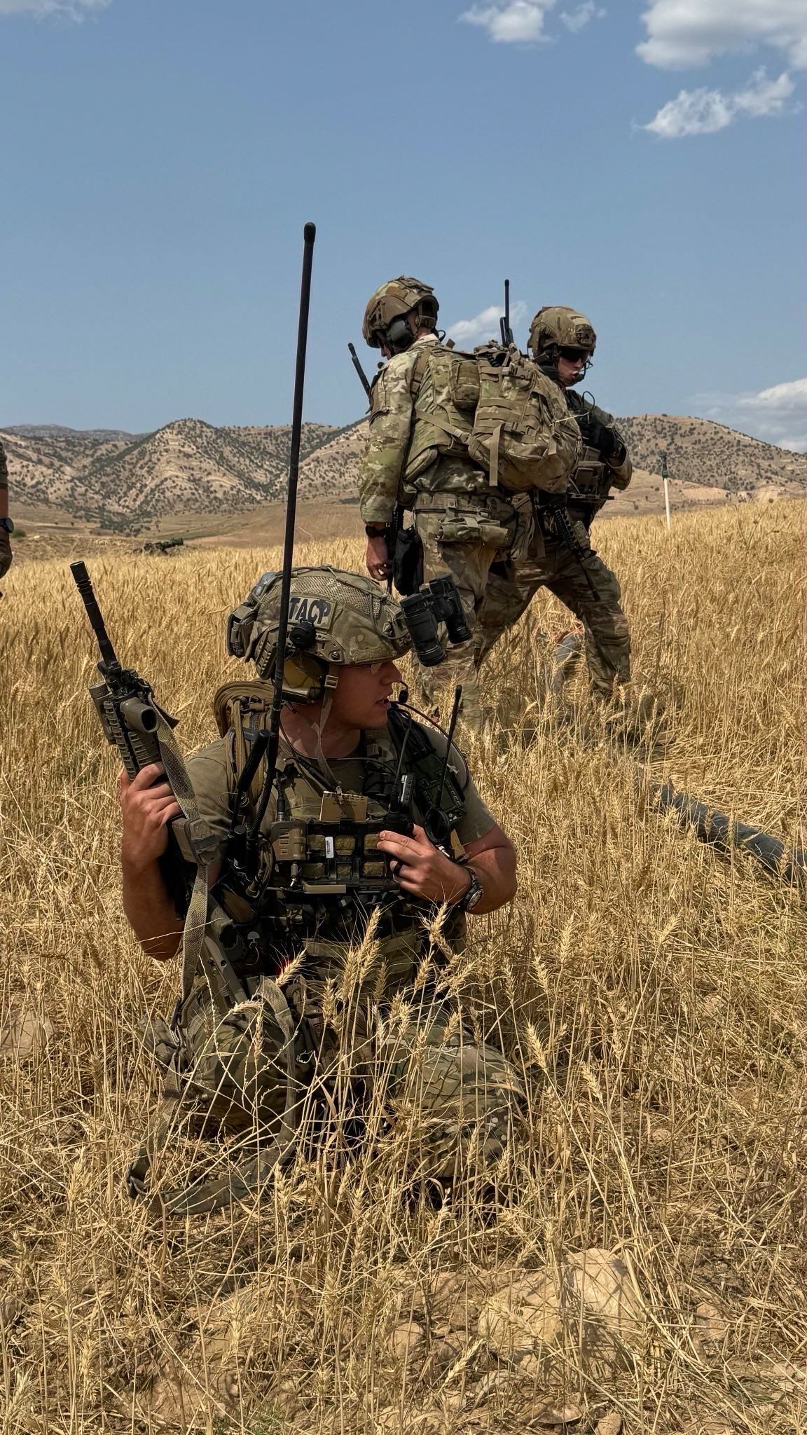 Military personnel in tactical gear move through dry grassland terrain with mountains visible in the background.