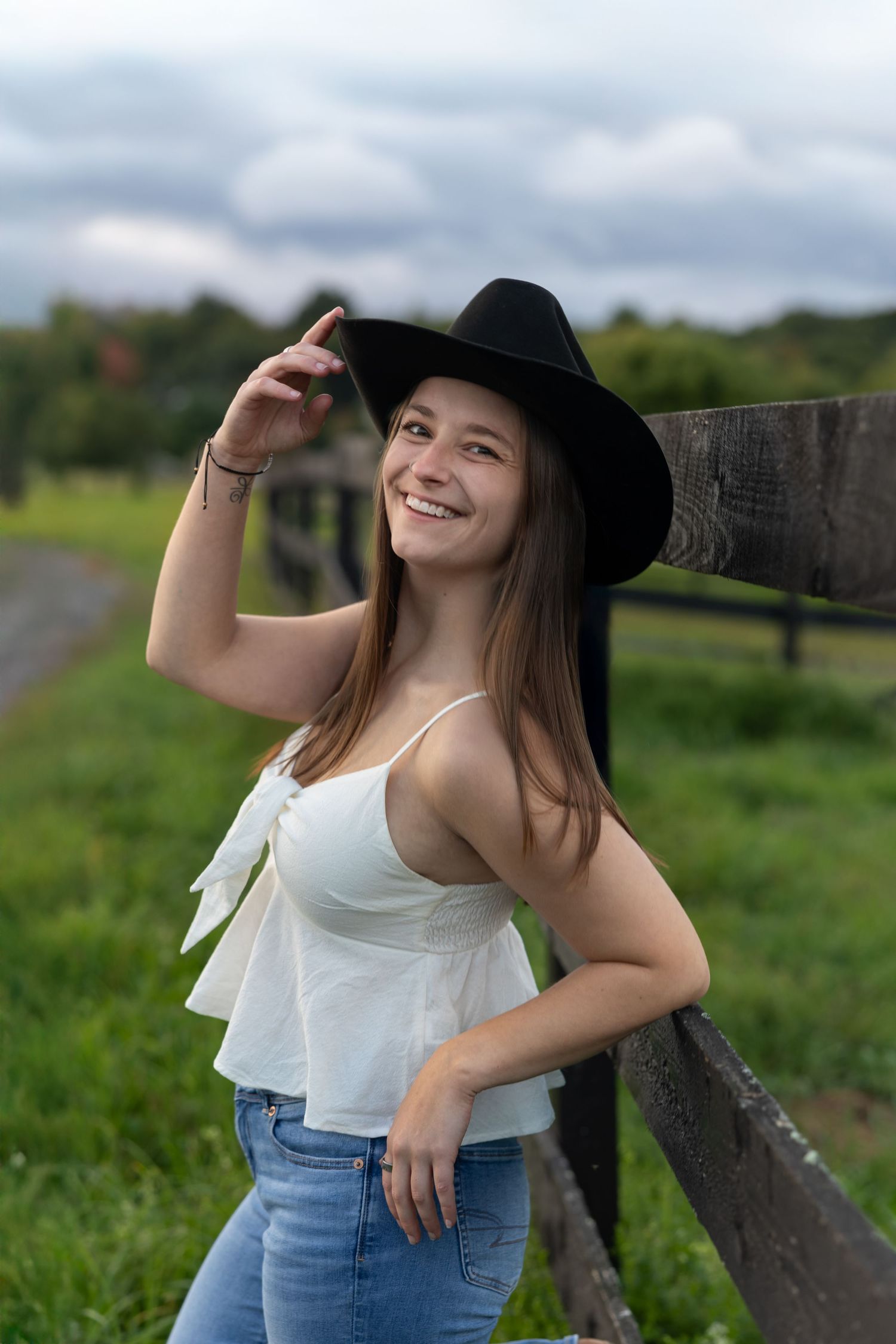 A person in a white top and jeans poses casually against a wooden fence in a rural setting on a sunny day.