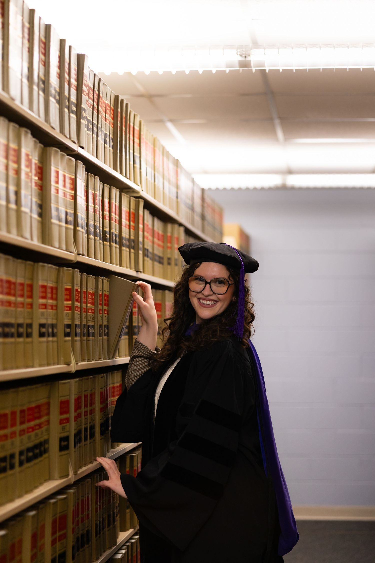 A graduate in academic regalia poses playfully between rows of library bookshelves.