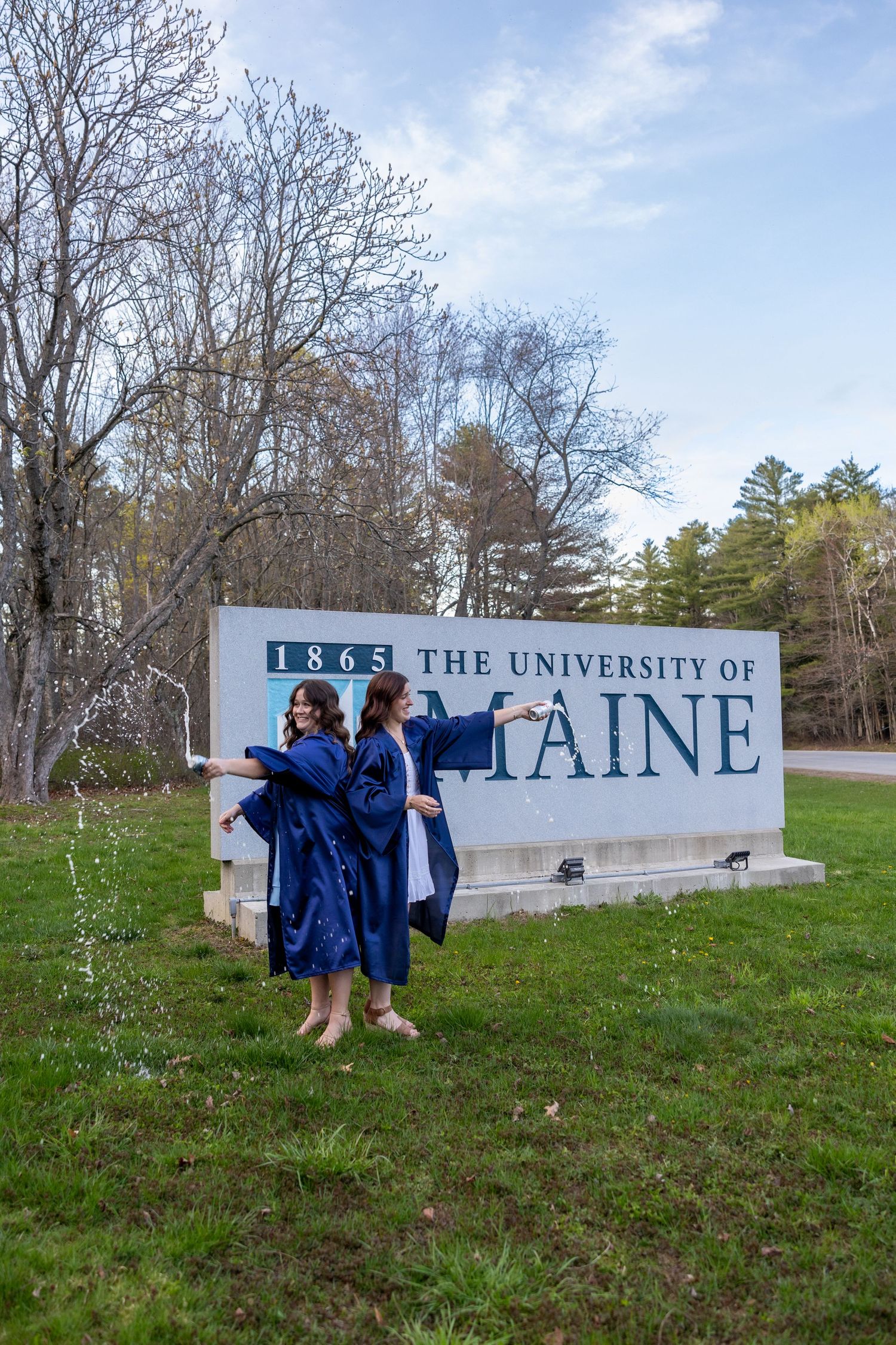 Graduates in blue robes celebrate near the University of Maine entrance sign on a spring day.