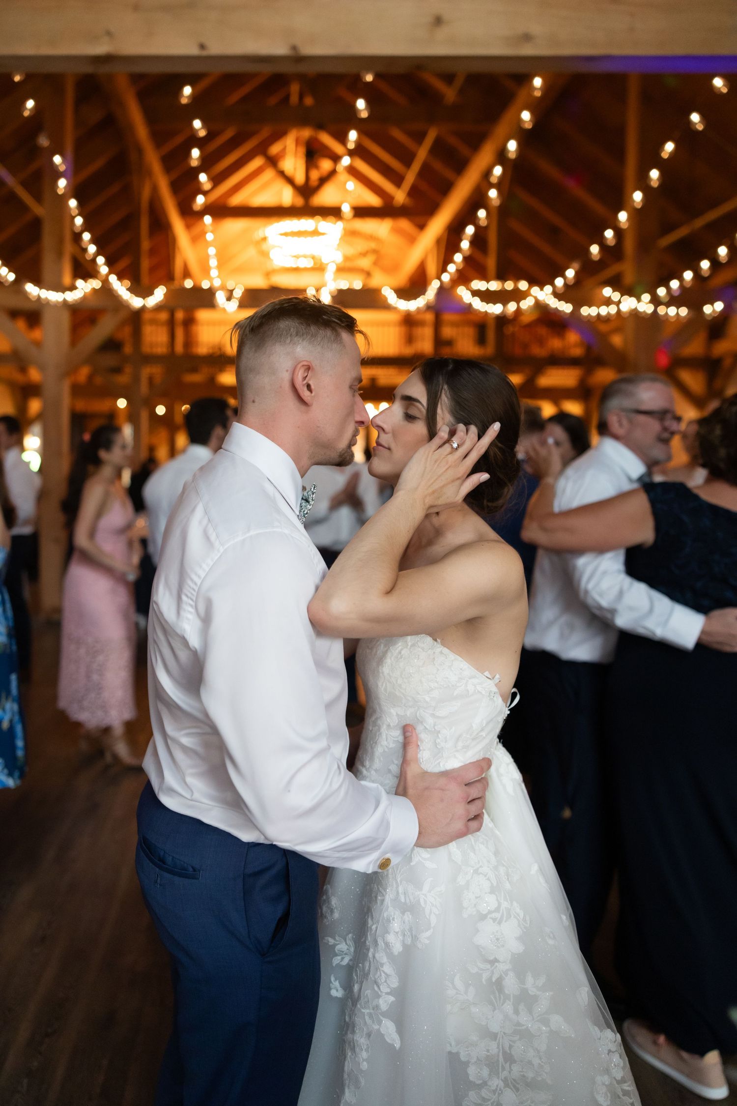 A couple shares their first dance at a rustic barn wedding reception with twinkling lights overhead.