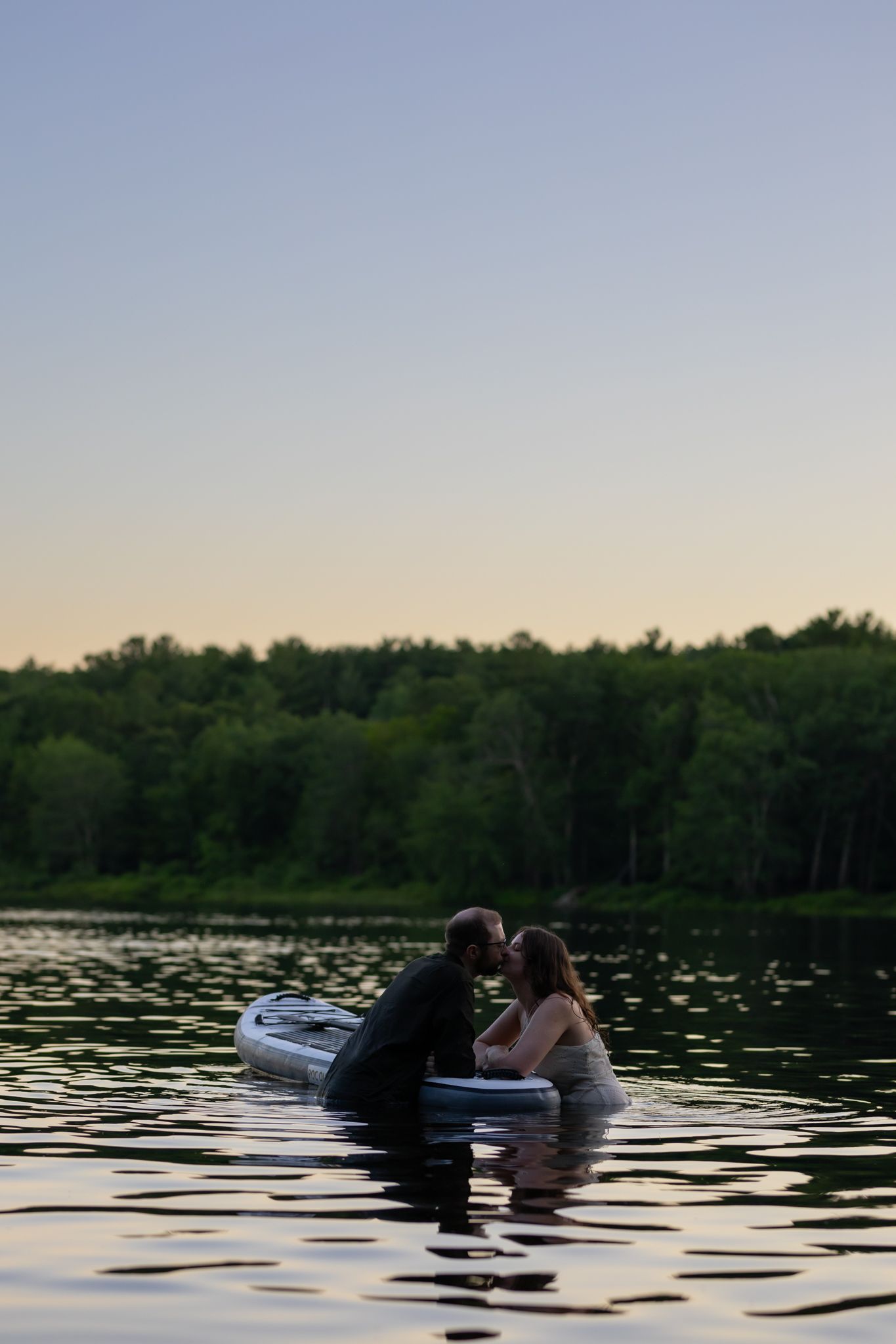Couple sharing a romantic kiss on a paddleboard in a calm lake at dusk, surrounded by lush green trees.