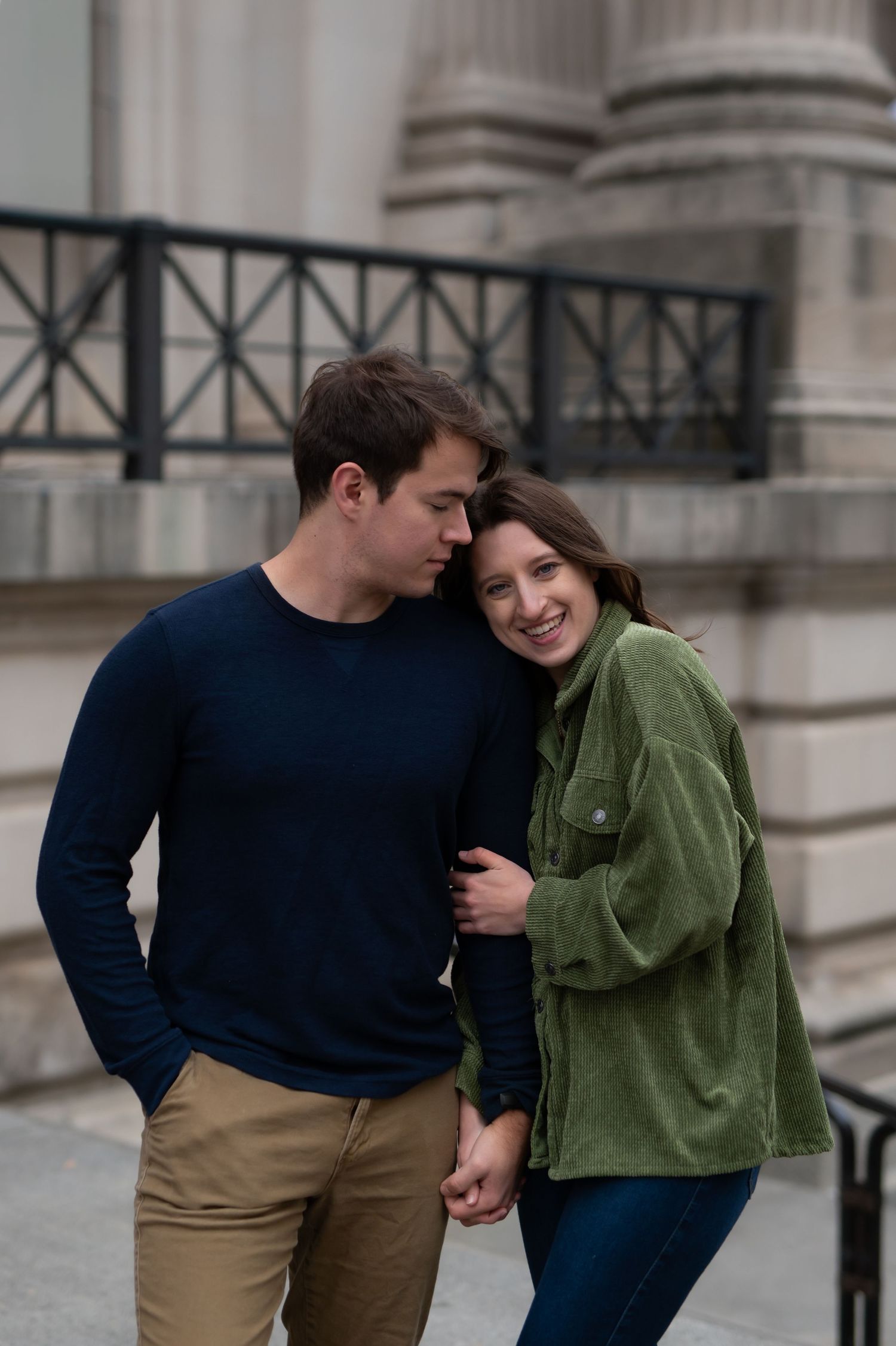 A couple shares an intimate moment together outside a building with classical columns, wearing casual sweaters.