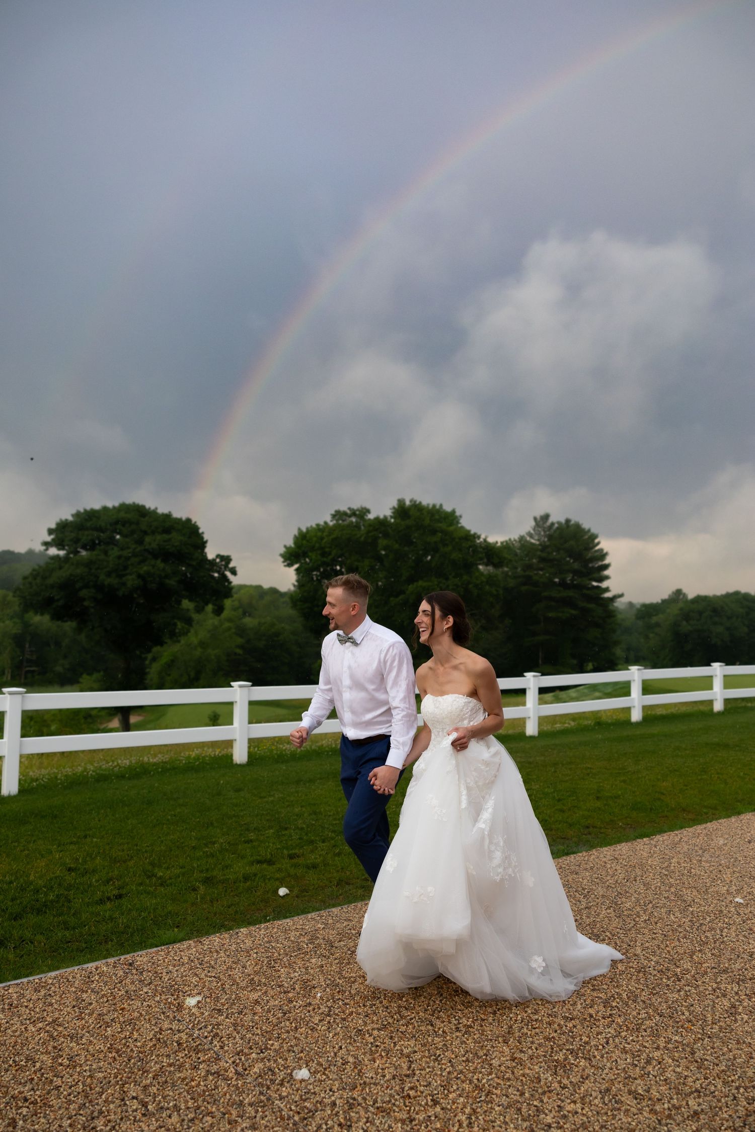 A couple in wedding attire runs along a gravel path next to a white fence with a dramatic stormy sky and rainbow above.
