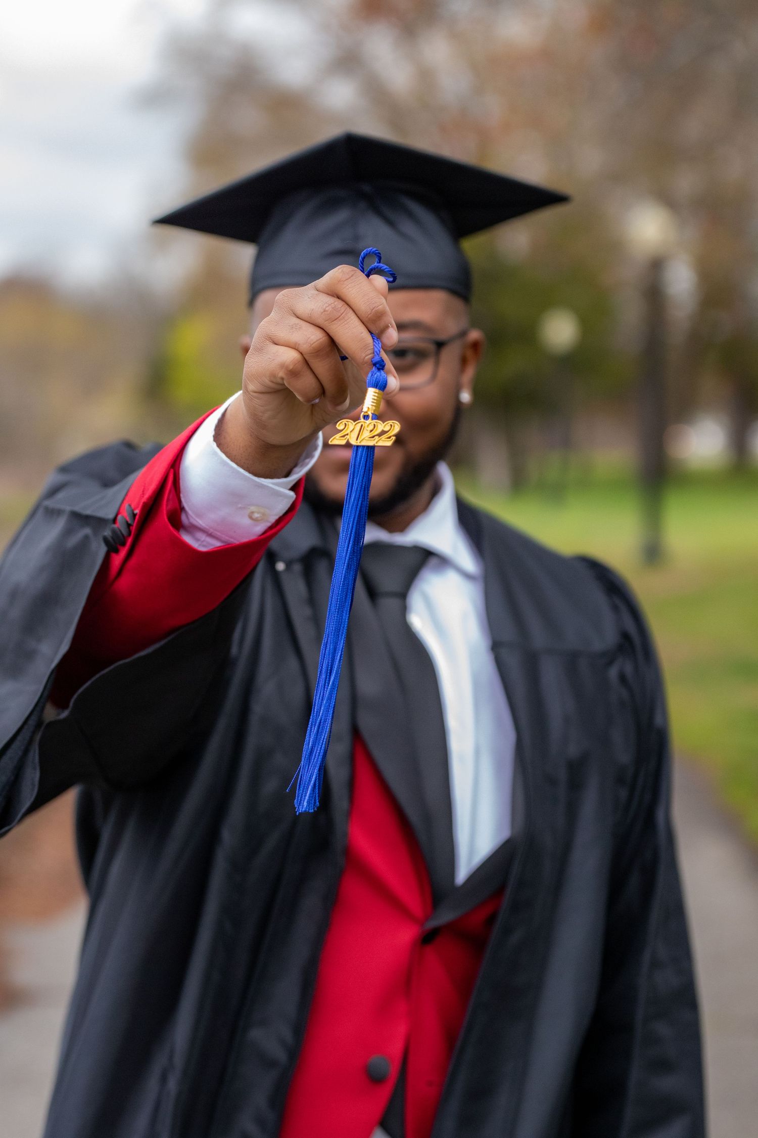 Graduate in cap and gown holds up a blue and gold graduation tassel against a blurred outdoor background.