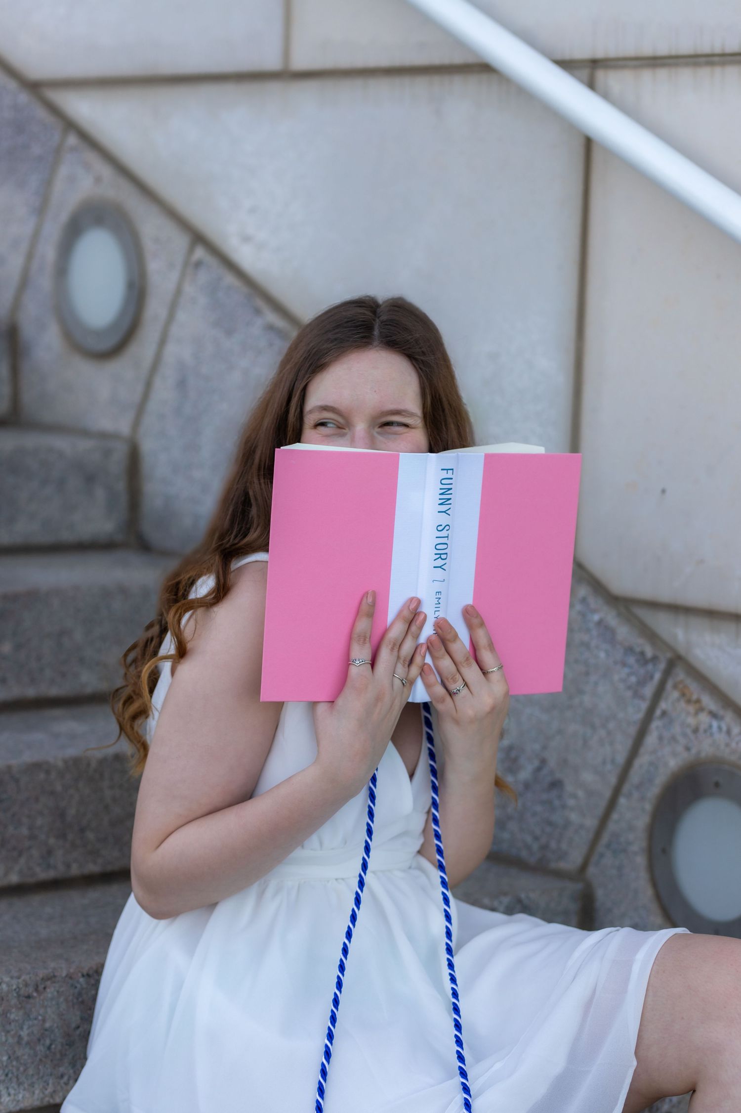 Person in white dress holds pink book in front of face while sitting on stone steps.