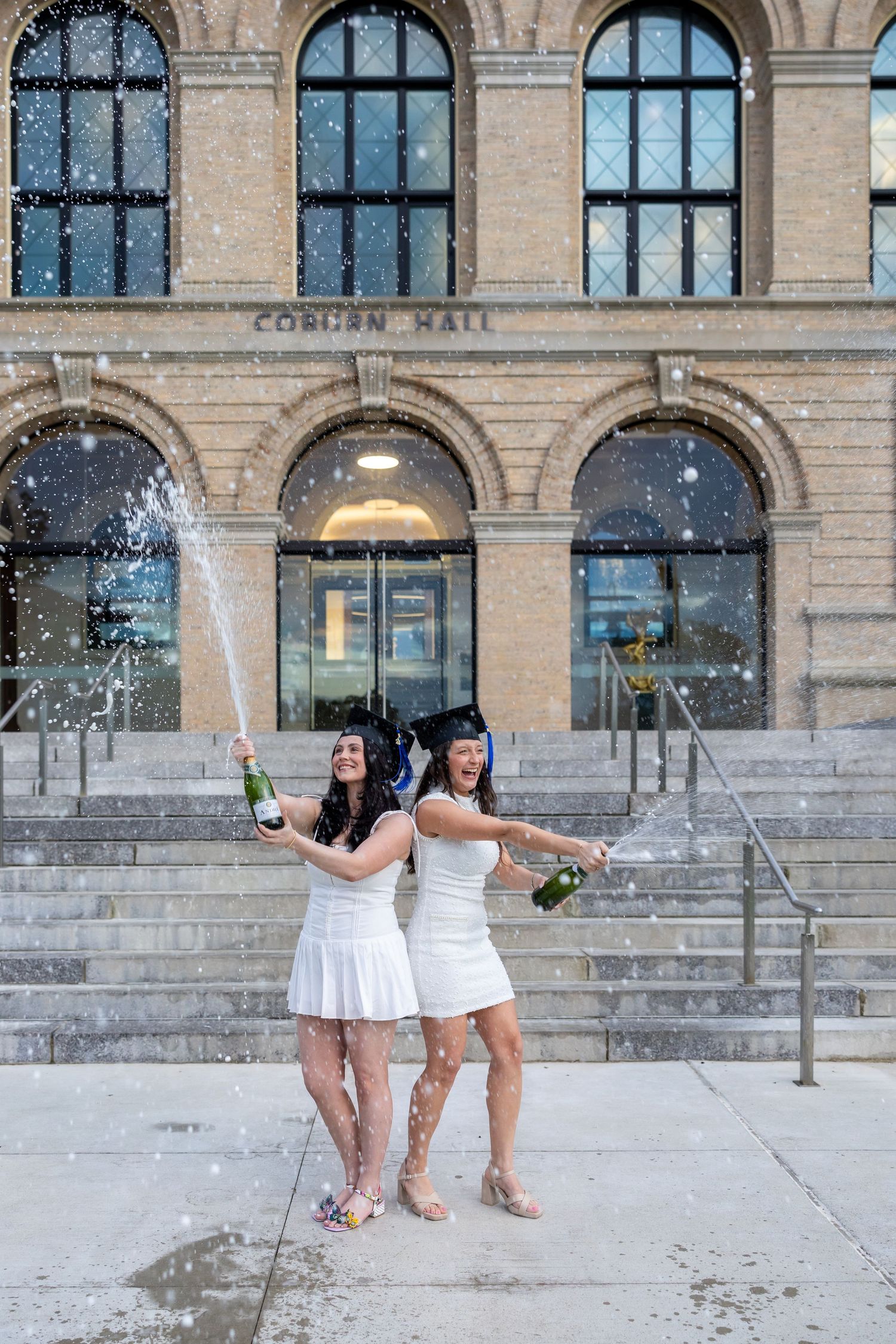 Two people in white dresses celebrate with champagne on grand stone steps of a historic building.