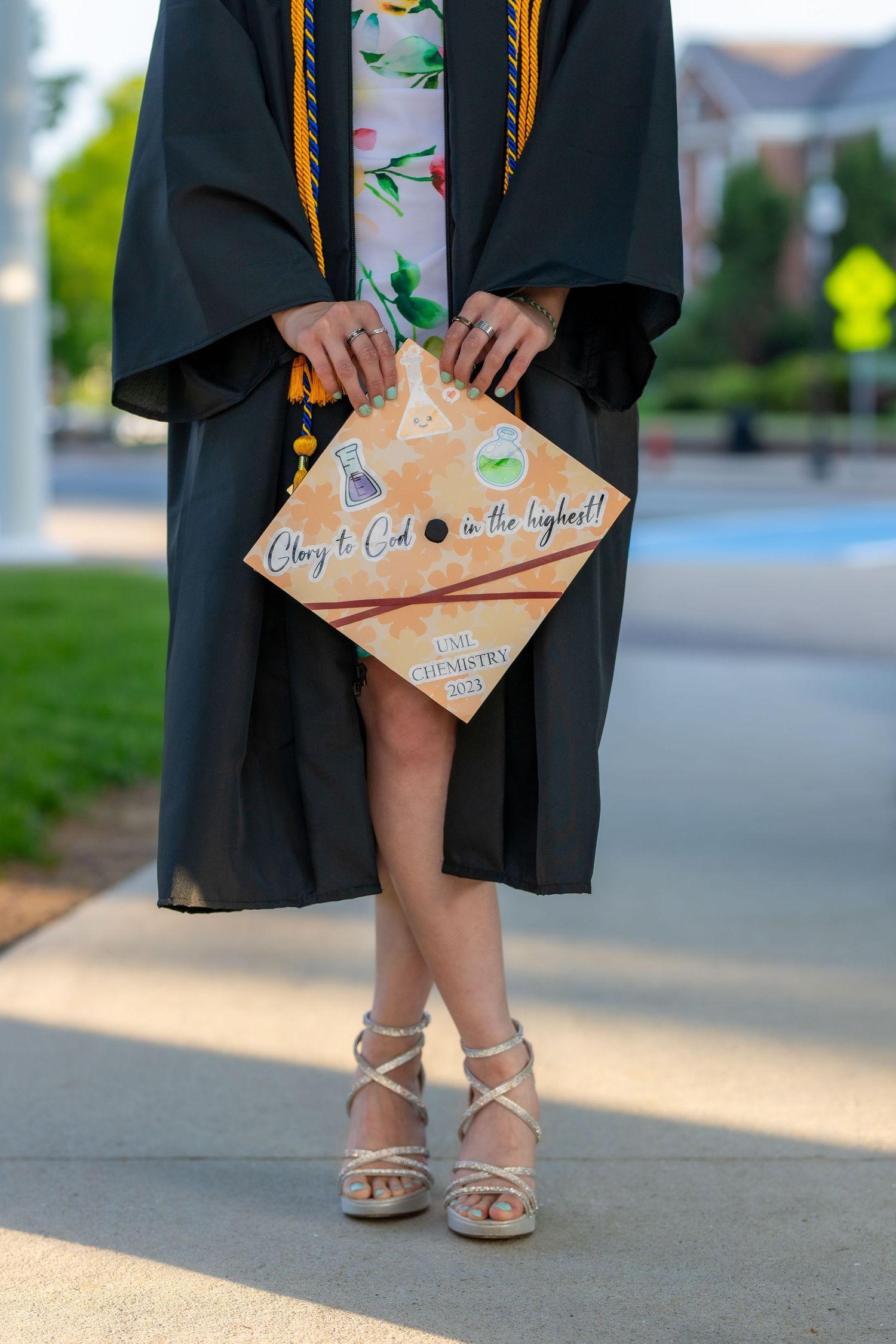 Graduate in black cap and gown holds decorated diploma holder while showing off strappy beige sandals.