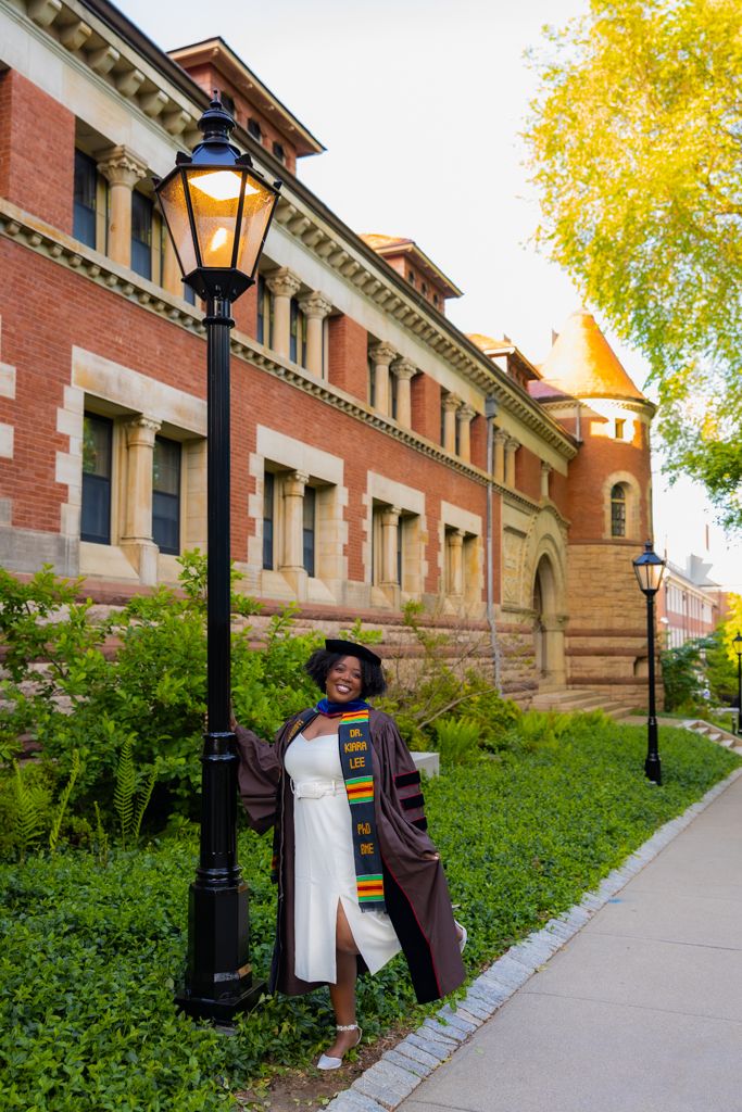 Graduate in academic regalia poses next to vintage lamp post outside historic brick university building in autumn.