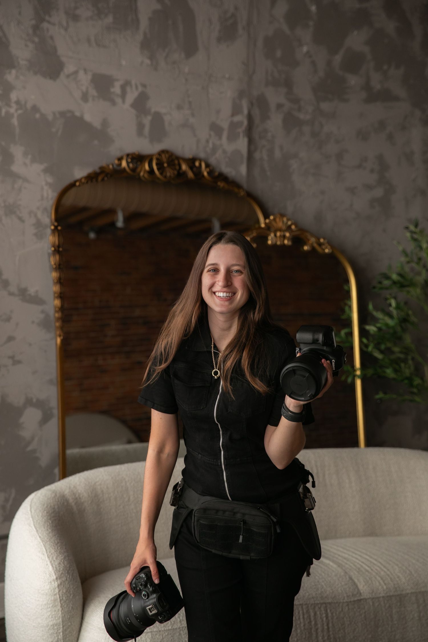 A photographer in all black attire sits casually on a white couch while holding professional camera equipment.