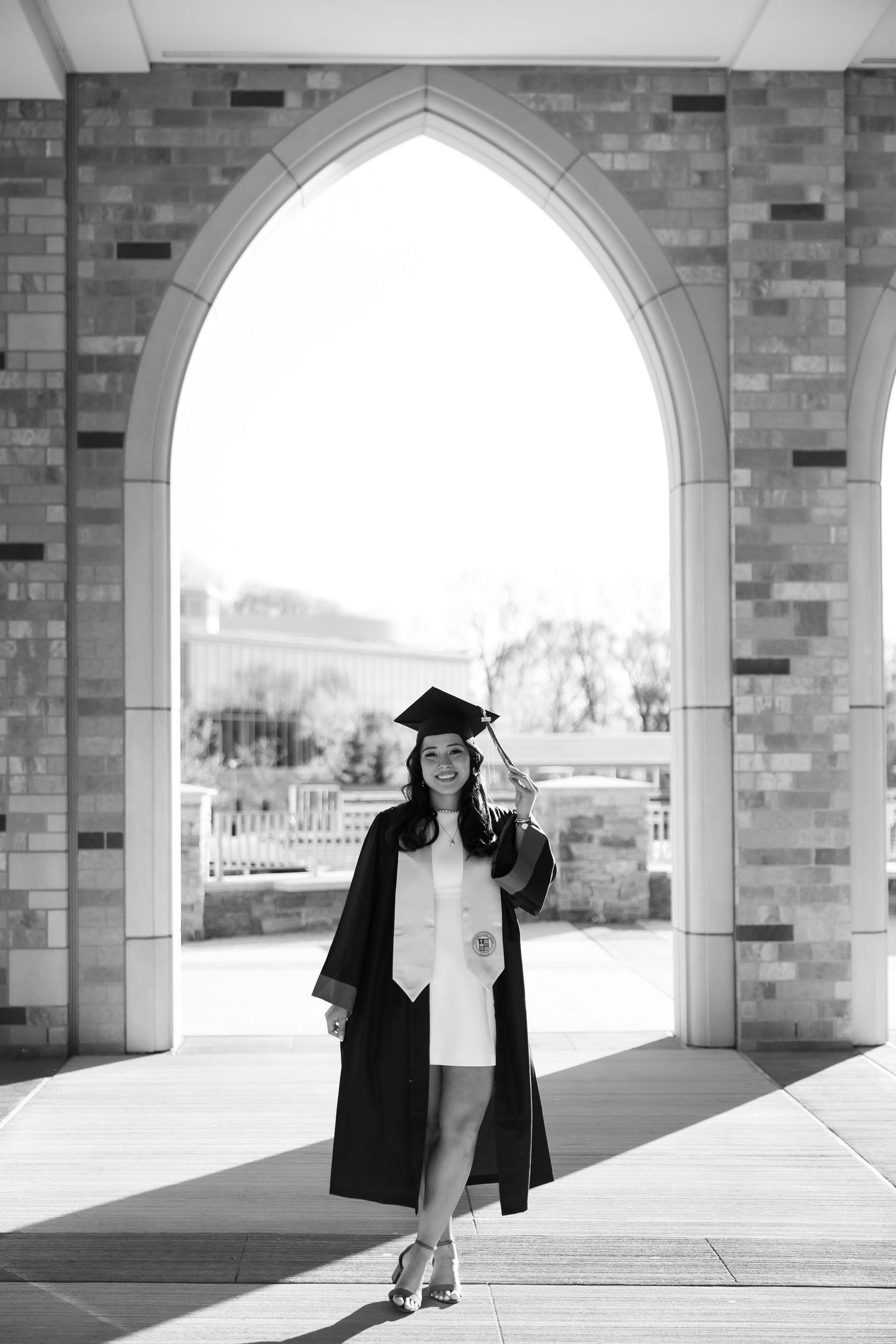 Black and white photo of a smiling graduate in cap and gown standing under an arched stone doorway.