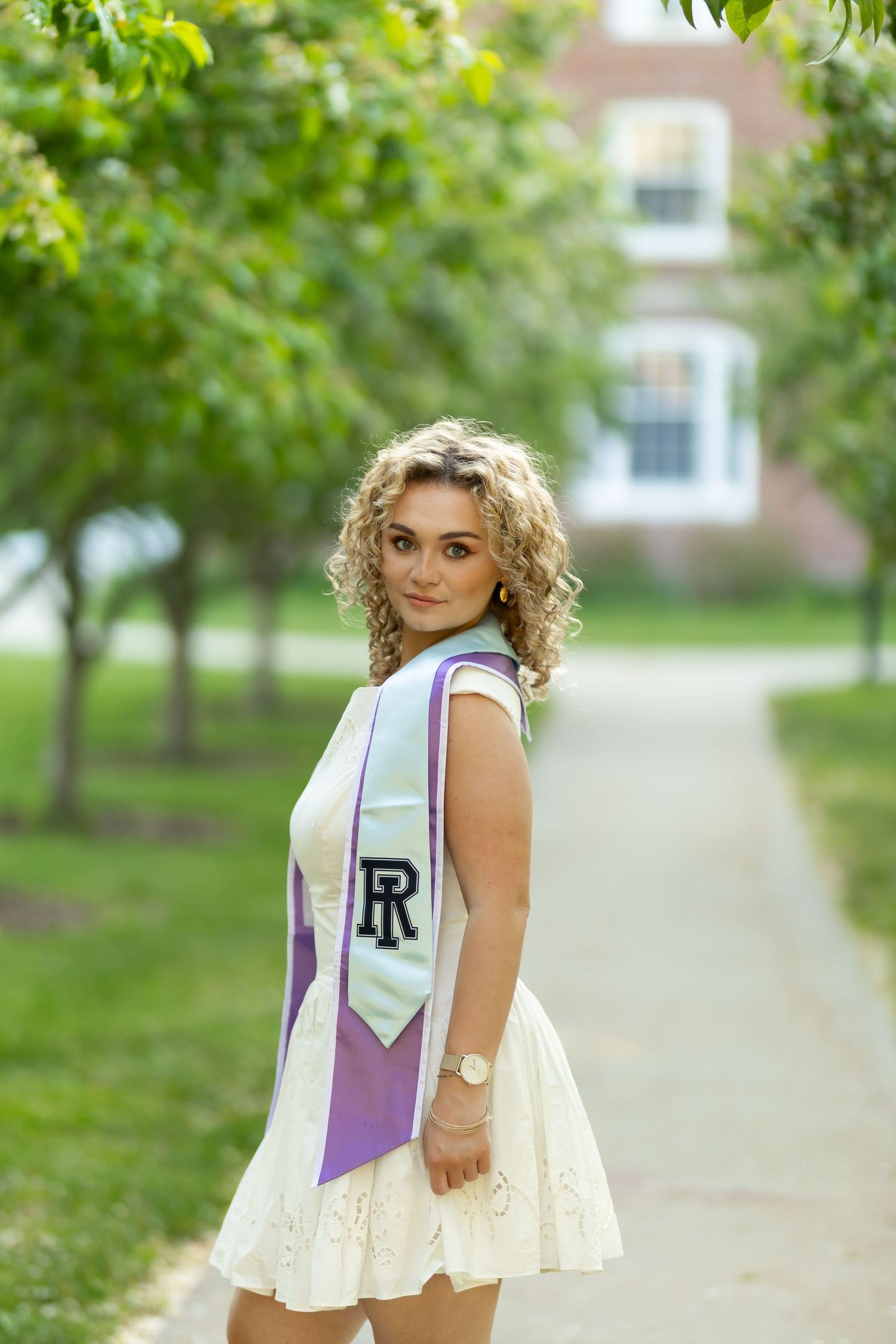 Young woman with curly blonde hair wearing a white dress and purple graduation sash poses on a tree-lined campus walkway.