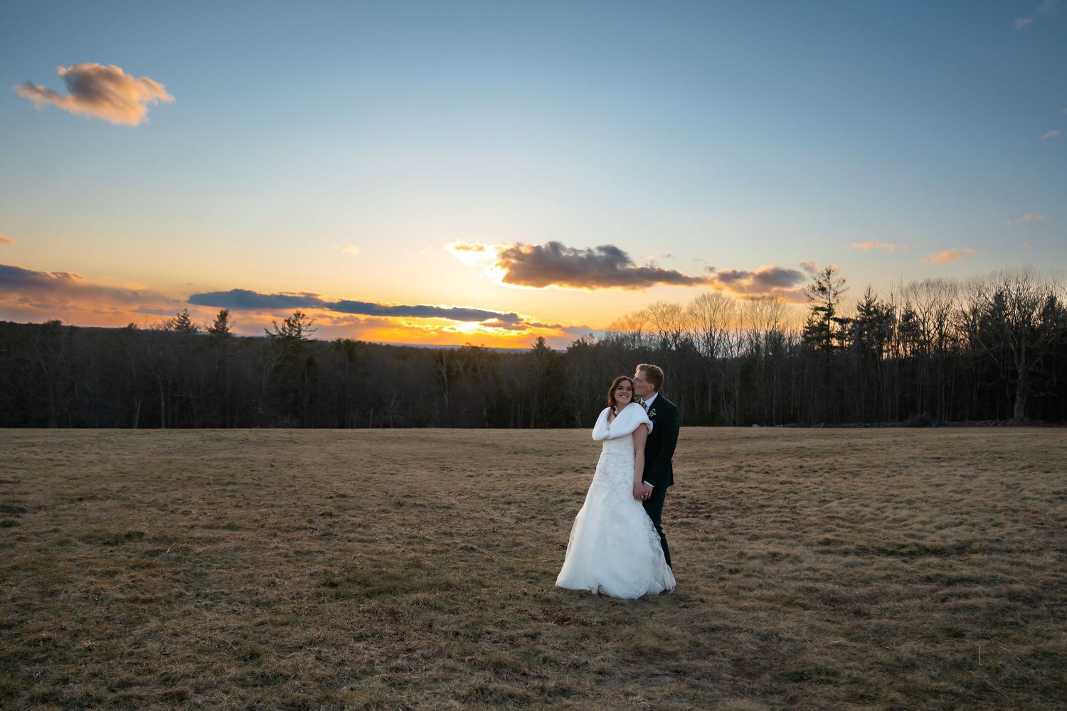 Bride and groom embrace in open field at golden sunset, trees silhouetted against vibrant sky on wedding day.