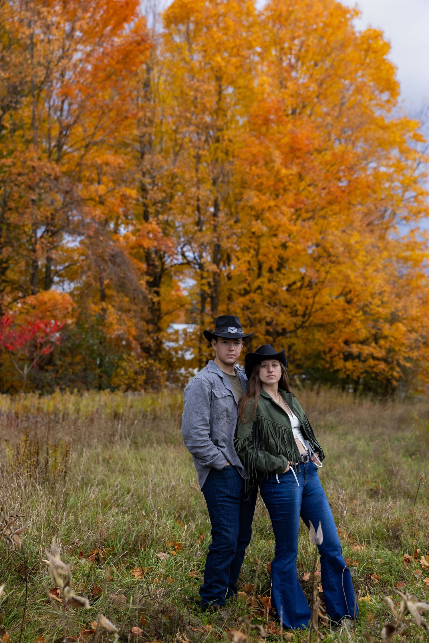 A couple stands together in a field against vibrant orange autumn trees on a fall day.