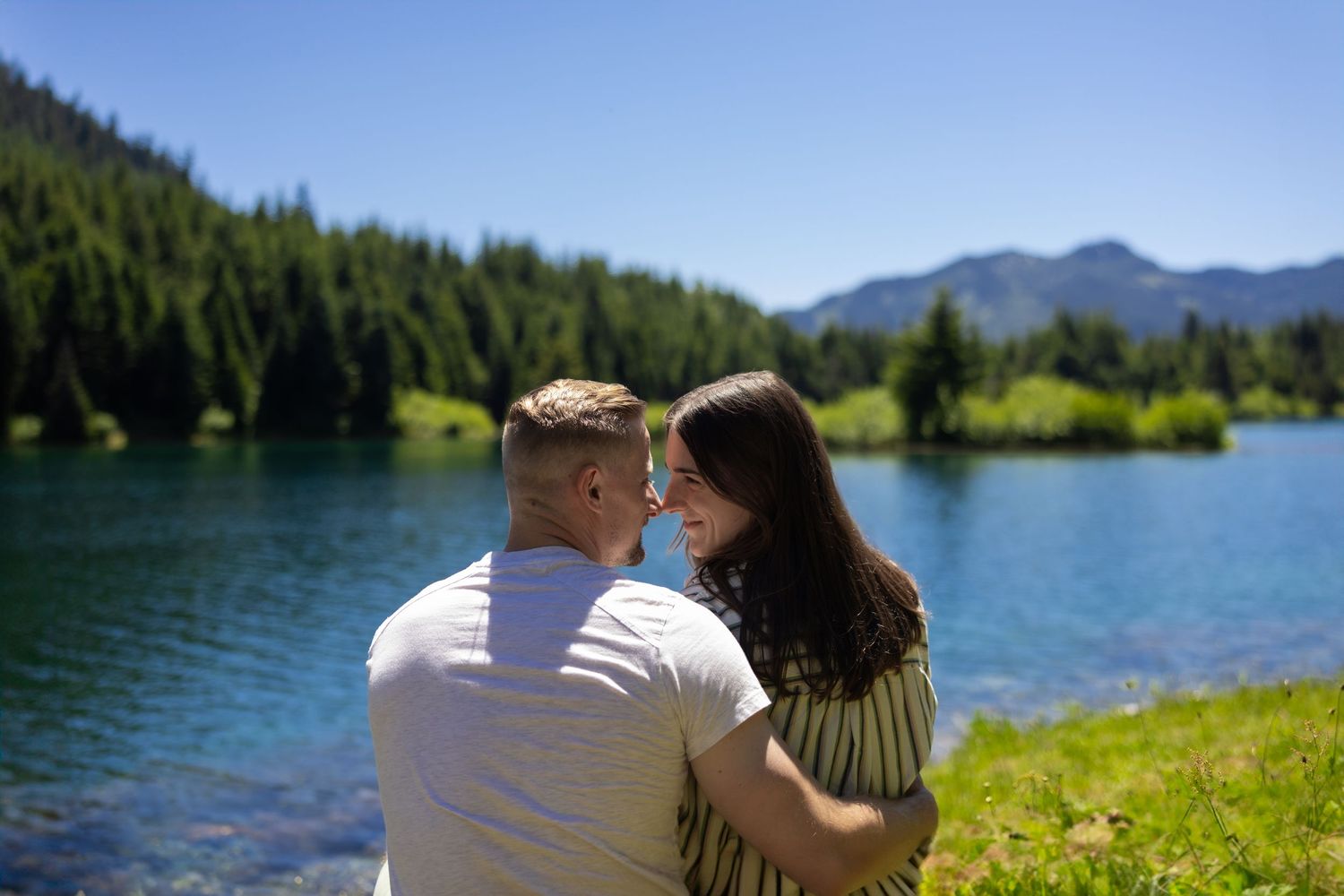 A couple sits together by a scenic mountain lake surrounded by evergreen forest on a sunny summer day.