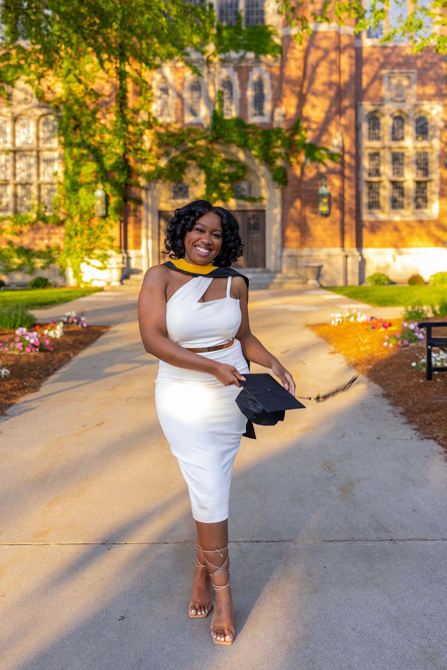 A person in a white sleeveless dress poses gracefully on a sunlit pathway with autumn trees and buildings in the background.