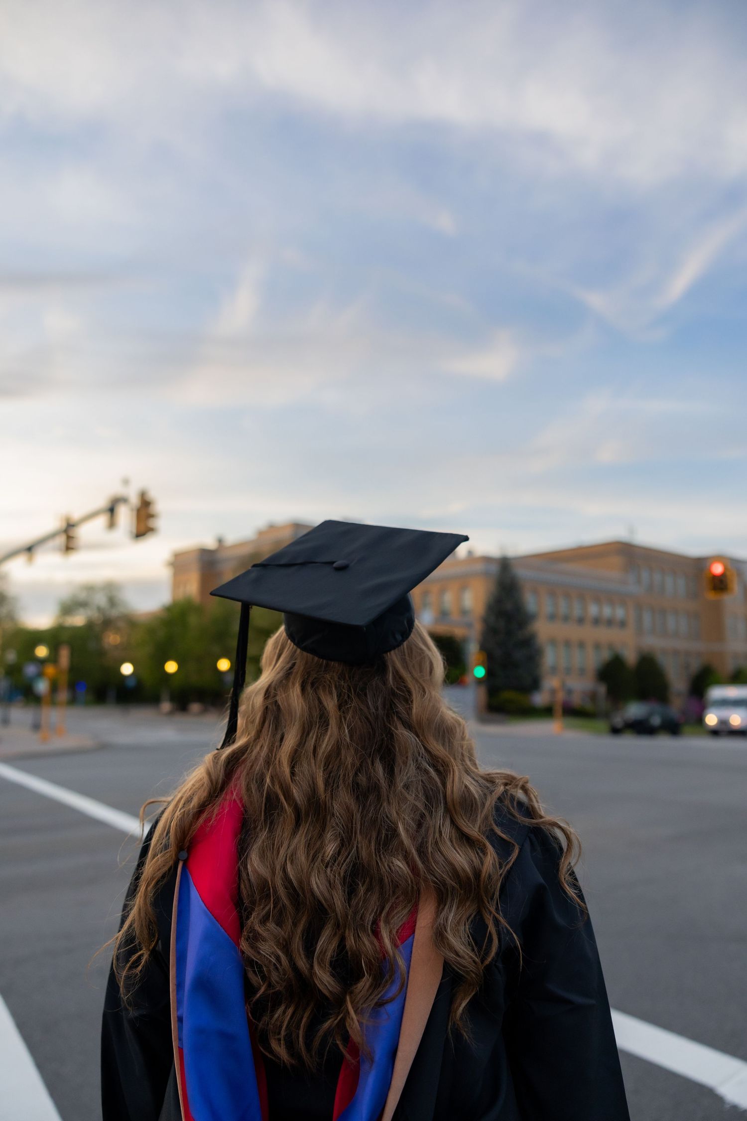 A graduate in cap and gown with long wavy hair stands in a parking lot on a sunny day.