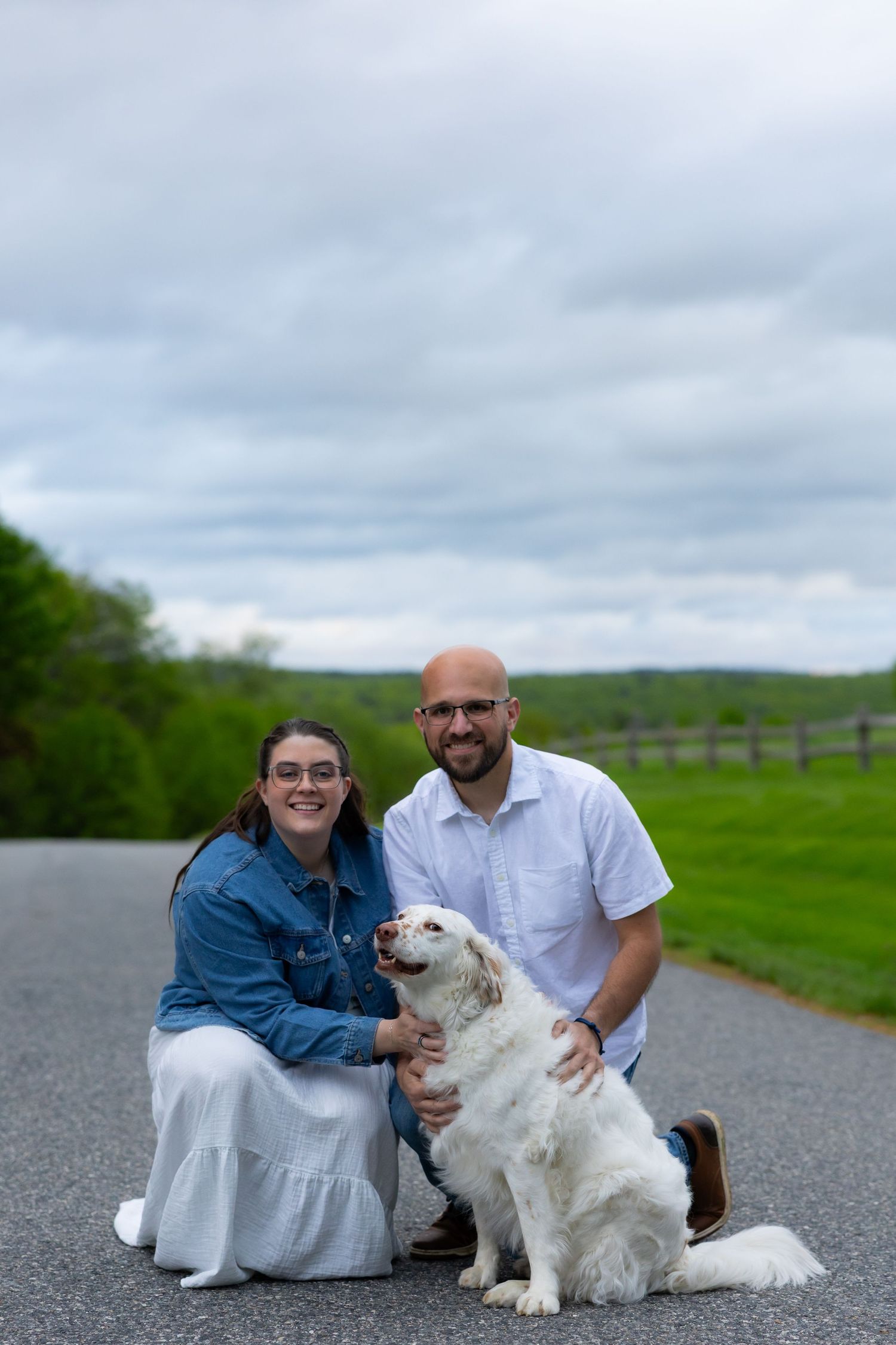 A couple poses with their fluffy white dog on a country road surrounded by green fields and cloudy skies.