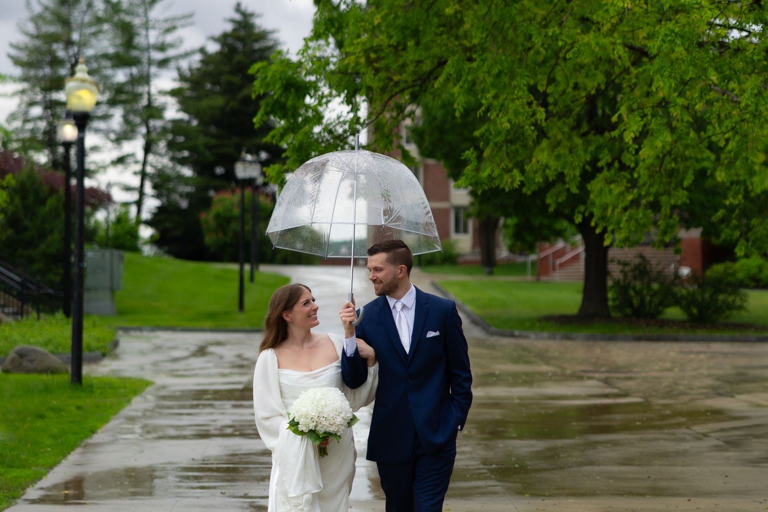 A couple in wedding attire walks together on a rainy day along a tree-lined path.