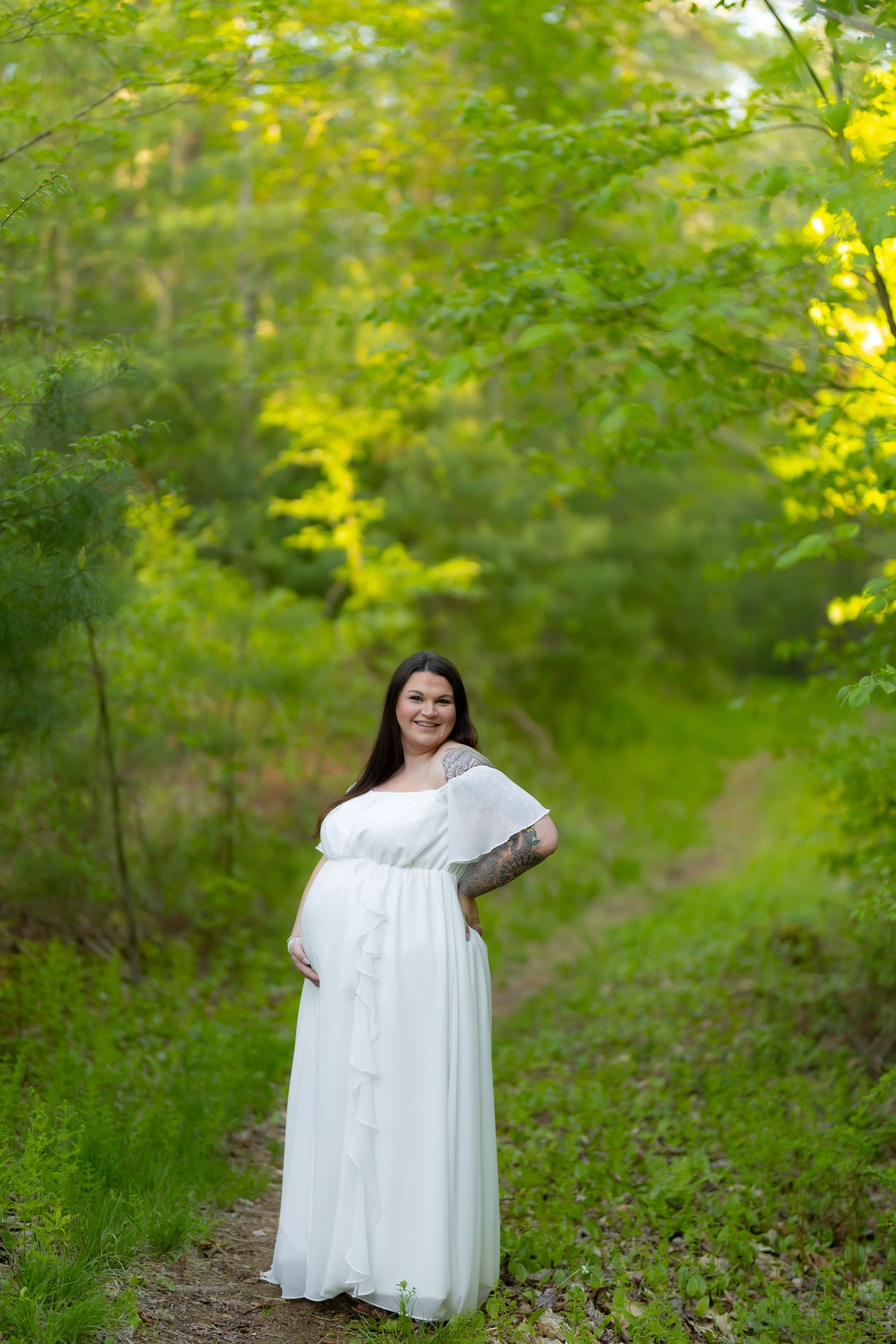 Pregnant woman in white dress standing on forest path surrounded by lush green trees during golden hour.