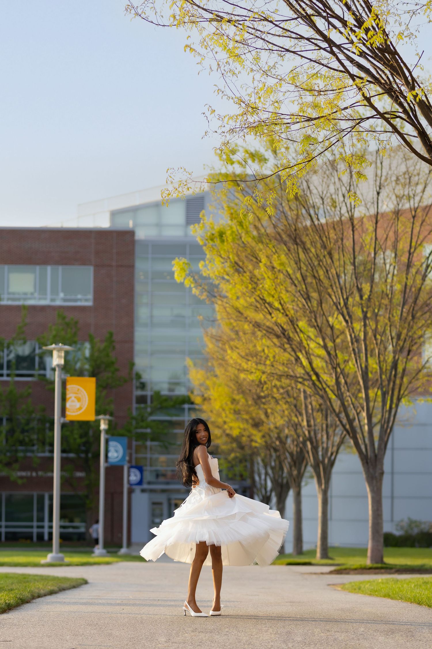Young woman in white tiered dress twirling on a campus pathway, surrounded by spring trees and brick buildings.