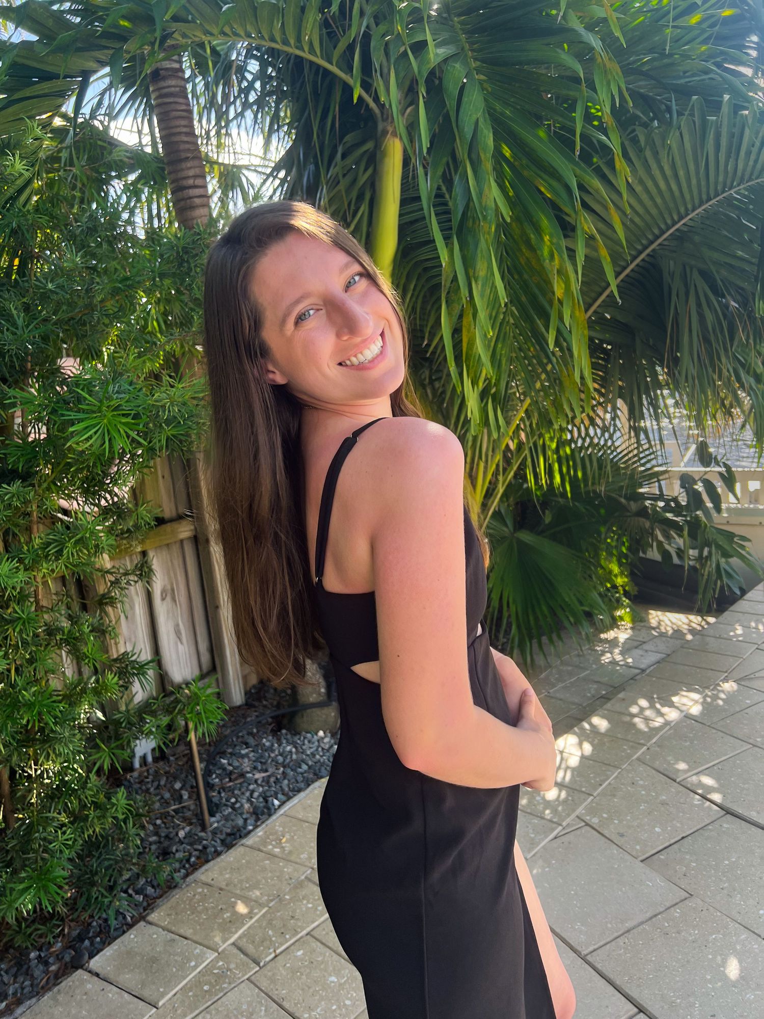 A person in a black dress poses against a backdrop of lush tropical plants and palm trees on a stone pathway.