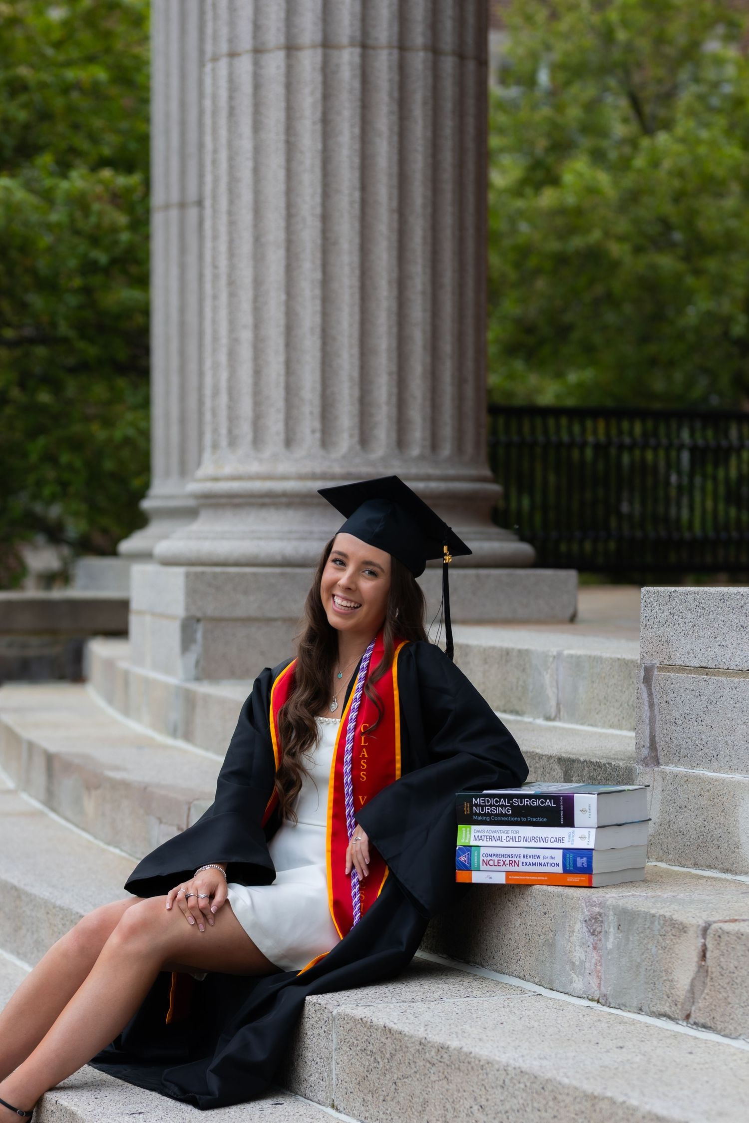 Graduate in cap and gown reclines on stone steps with textbooks beside classical columns during graduation photoshoot.