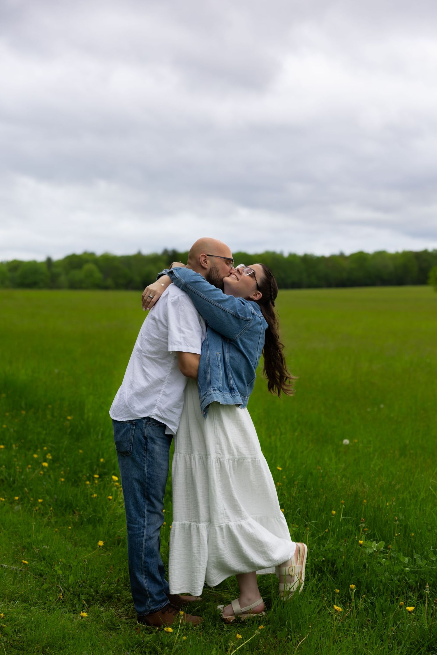 A couple embraces lovingly in a lush green meadow with wildflowers under an overcast sky.