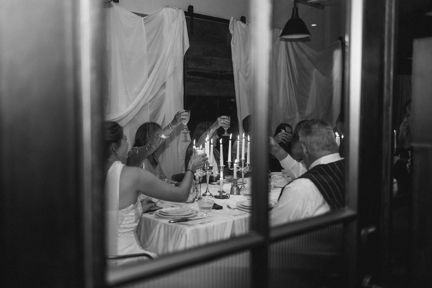Candlelit dinner reception viewed through a window with elegant table settings and white linens.