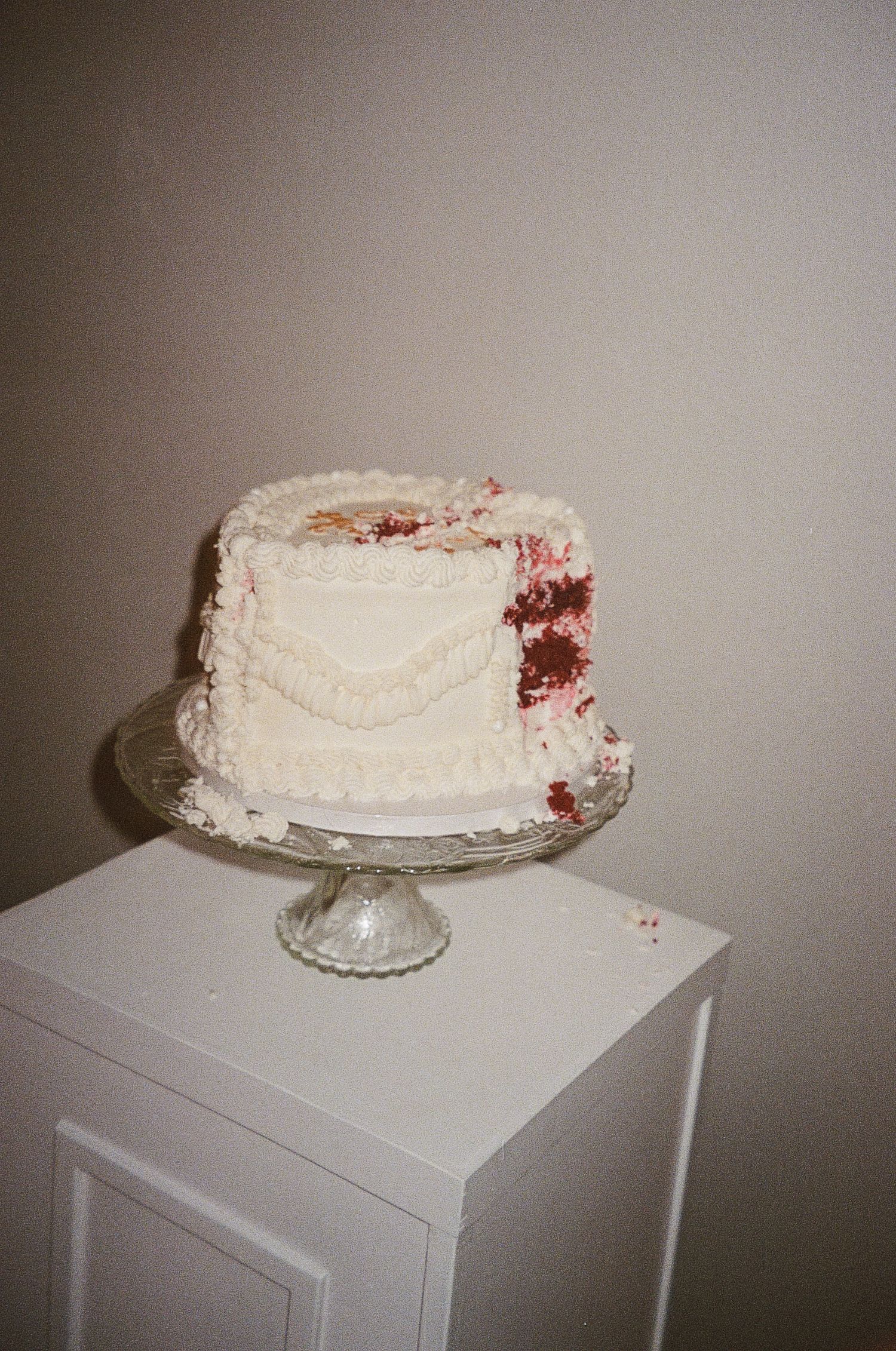 A white frosted cake with red accents displayed on a glass cake stand. Film Photograph.