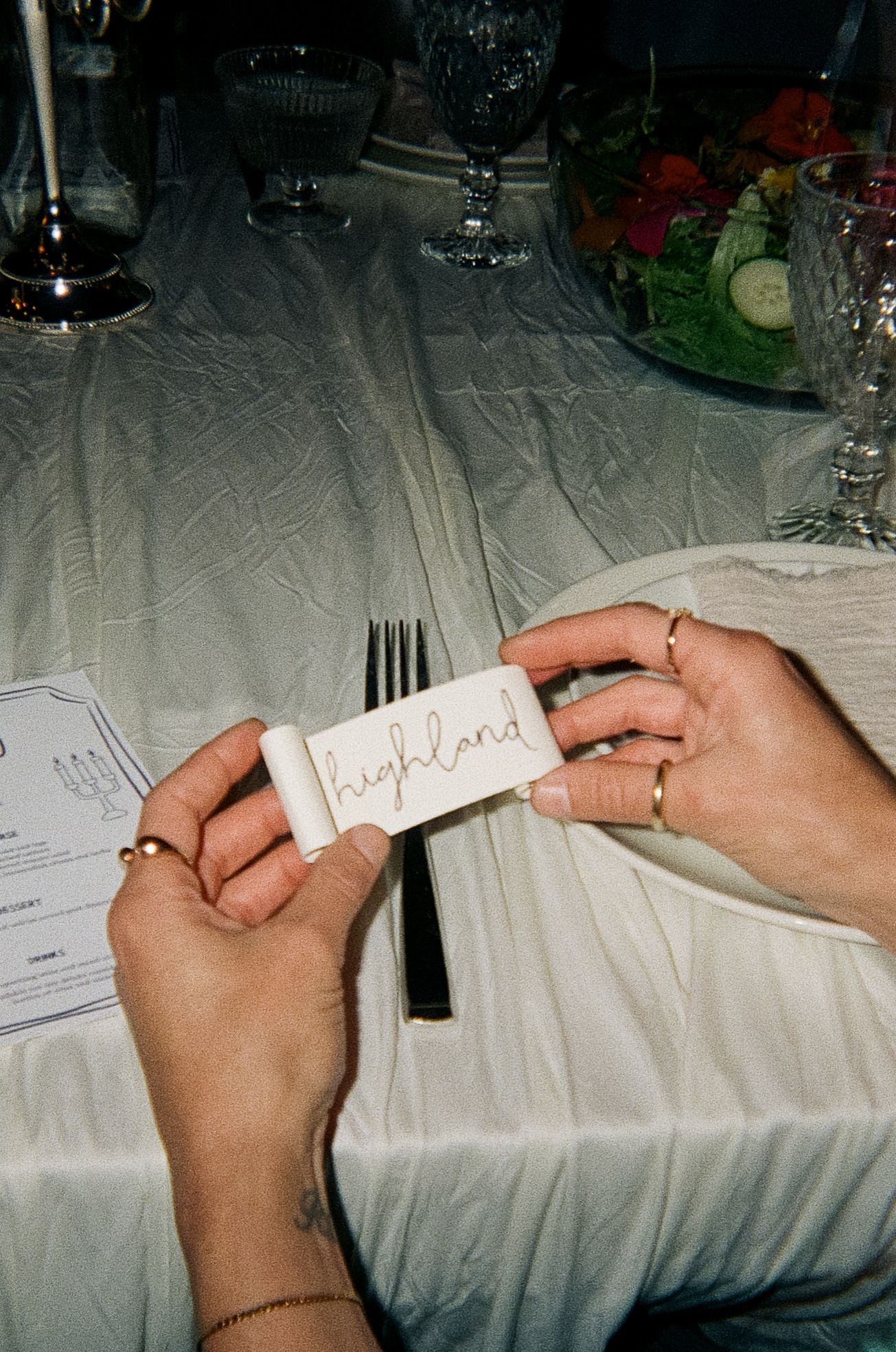 Hands holding a handwritten place card at an elegant table setting with wine glasses and linens. Film Photograph.