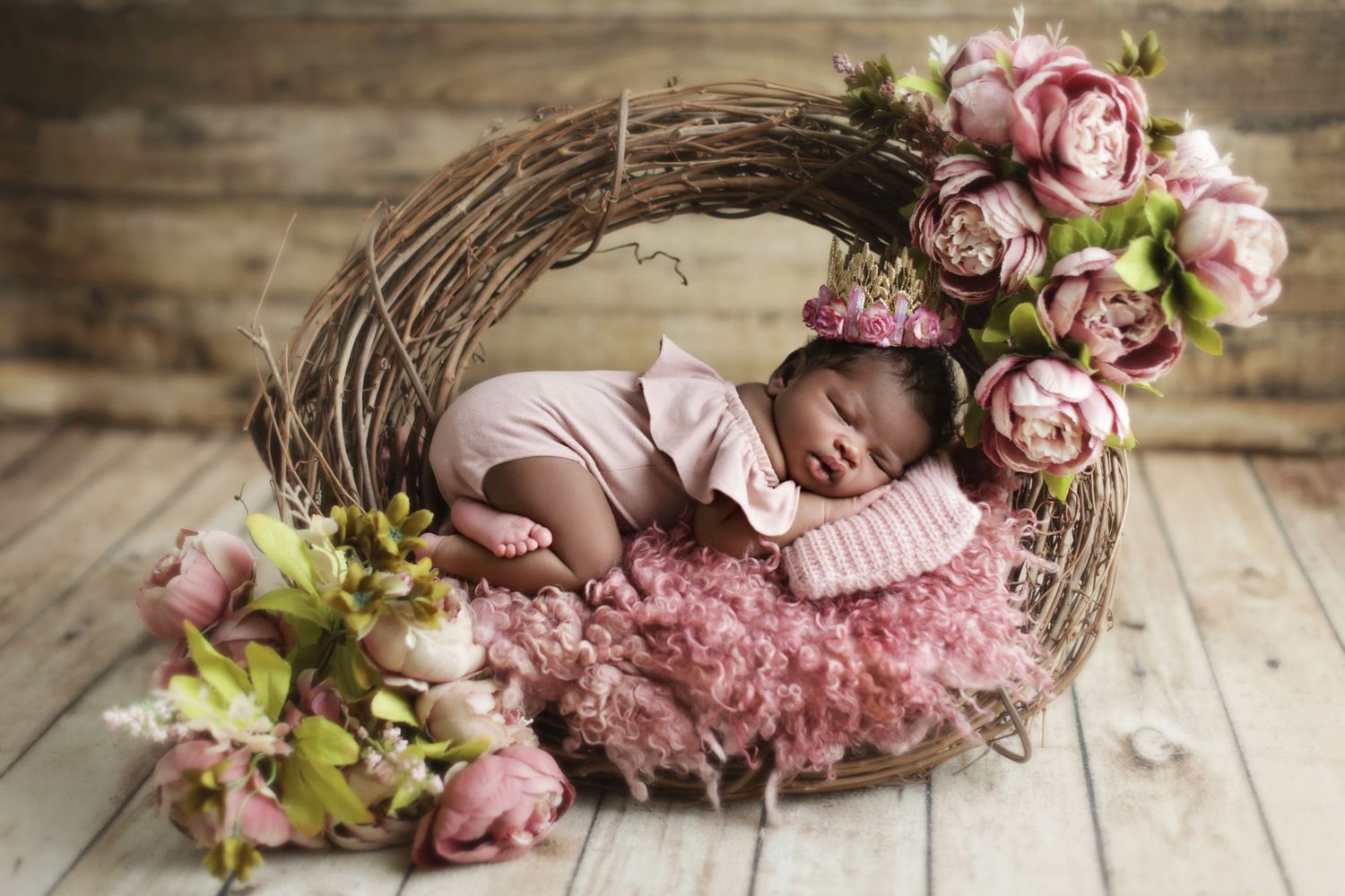 Newborn baby sleeping peacefully in a rustic twig nest with pink flowers and fluffy pink bedding.