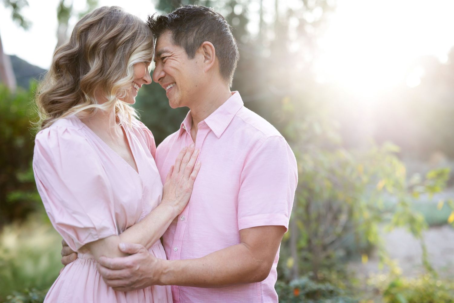 A happy couple in matching pink outfits embrace and smile at each other in a sunlit garden.