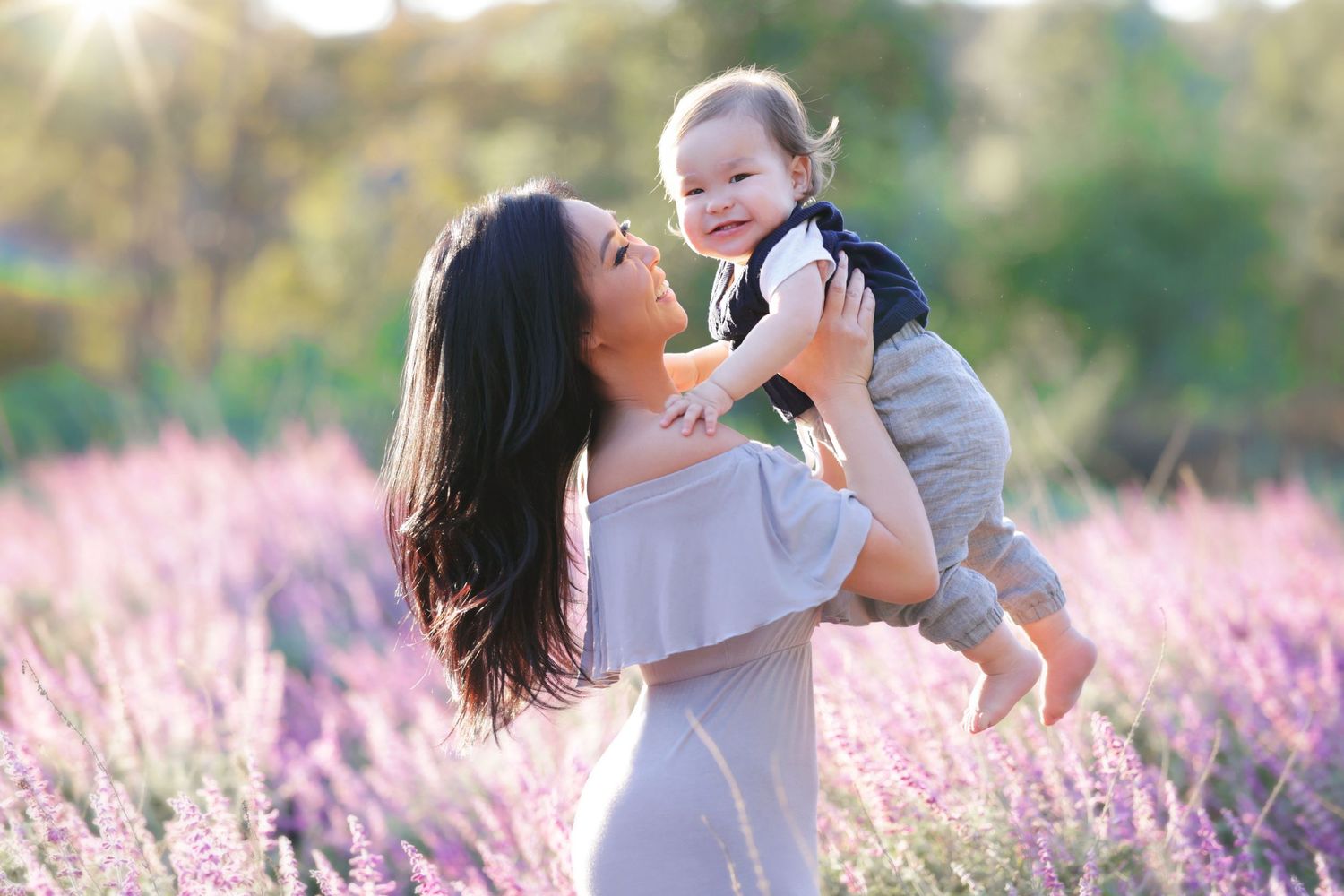 Woman holding smiling baby in lavender field with sunlight, joyful mother-child moment outdoors.