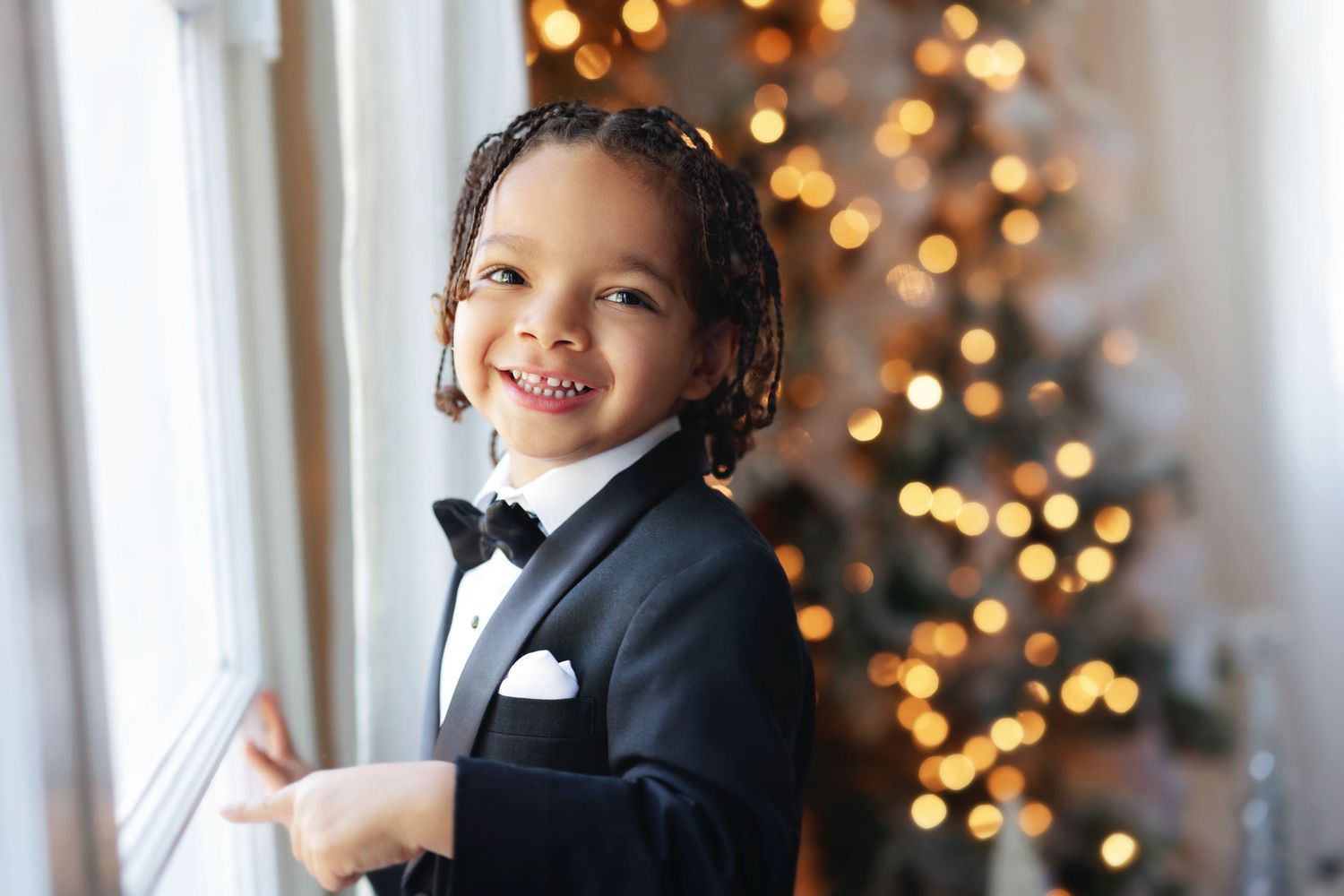 Smiling young boy in a tuxedo with bow tie standing by a window with a decorated Christmas tree.