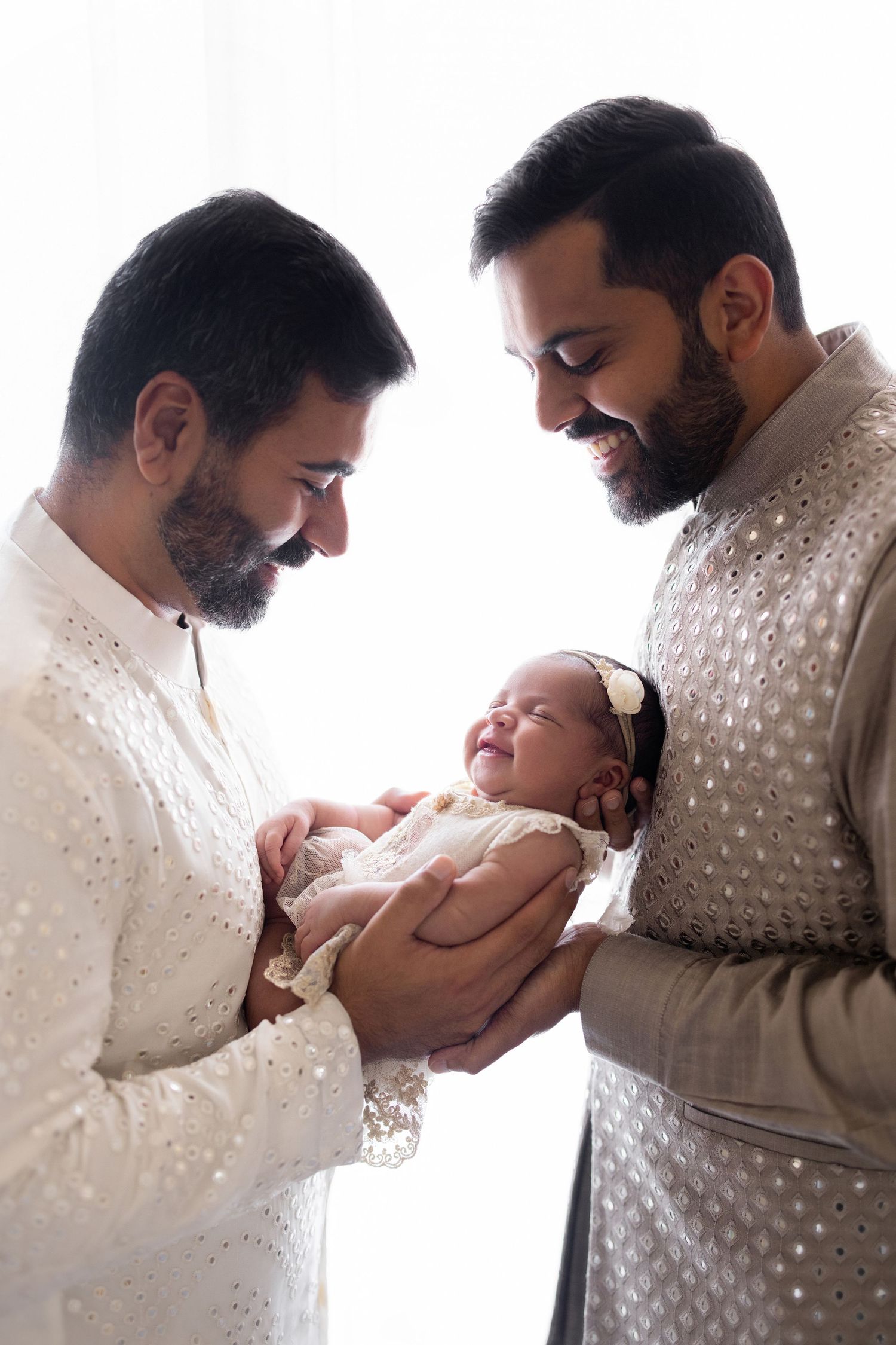 Two bearded men in traditional Indian attire lovingly hold a smiling newborn baby girl in a bright studio setting.
