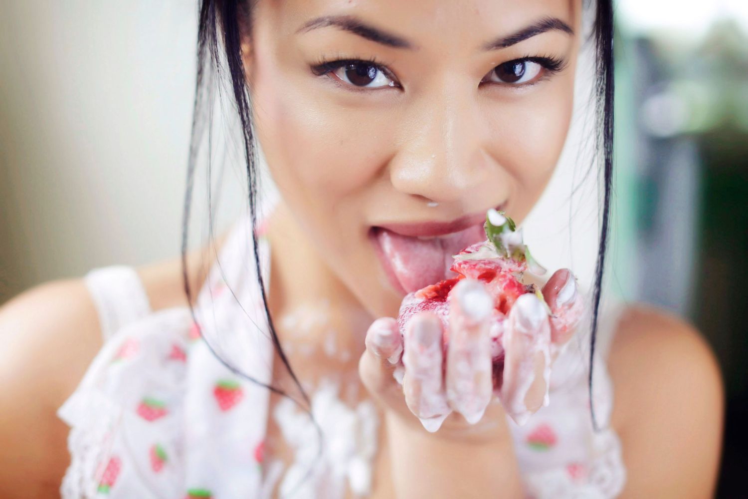 Young Asian woman licks cream from a strawberry dessert in a playful and expressive close-up portrait.