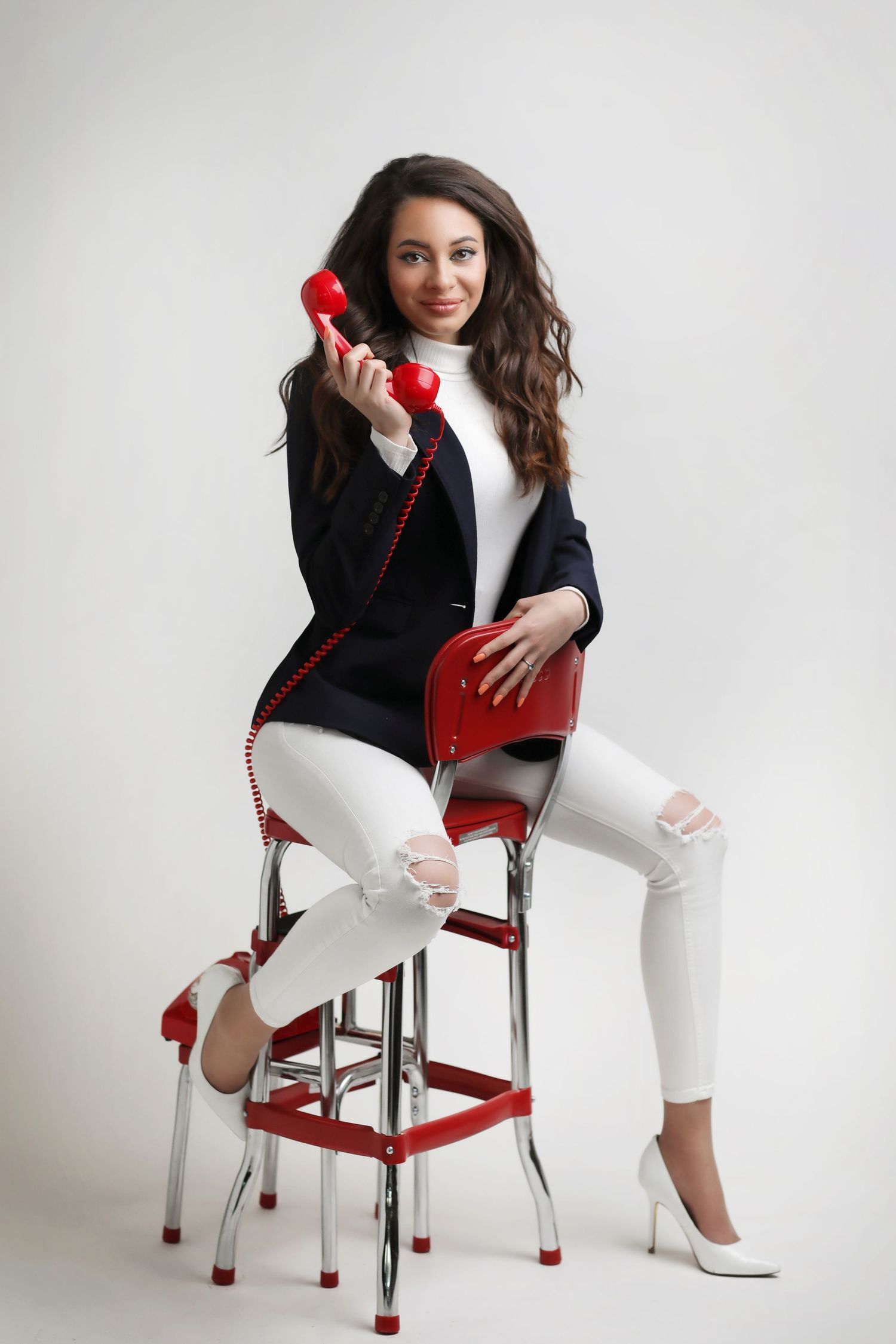 Young woman in white jeans and blazer sitting on red bar stool holding a vintage red telephone handset.