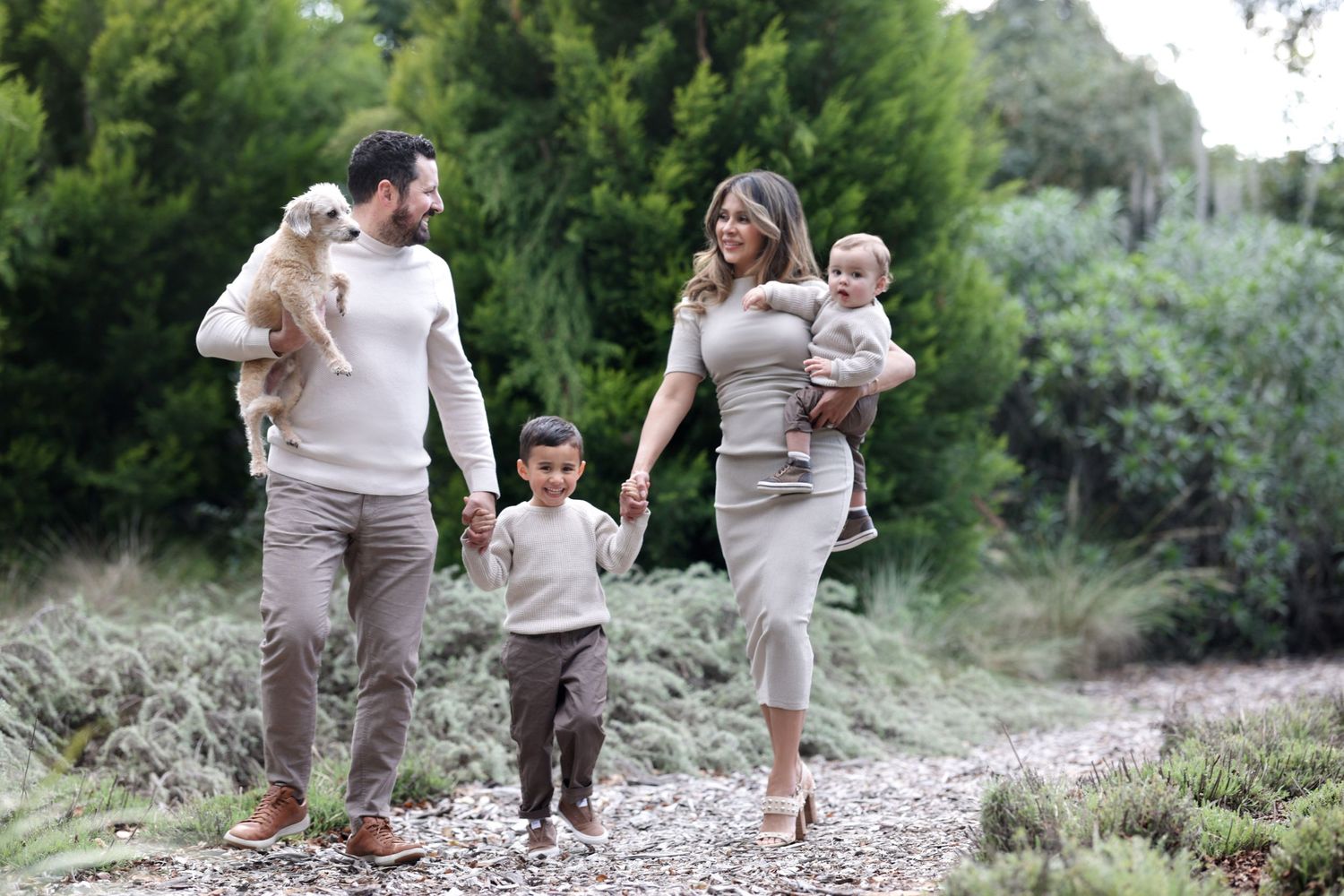 Smiling family with a toddler and baby strolling along a peaceful garden path with a dog.